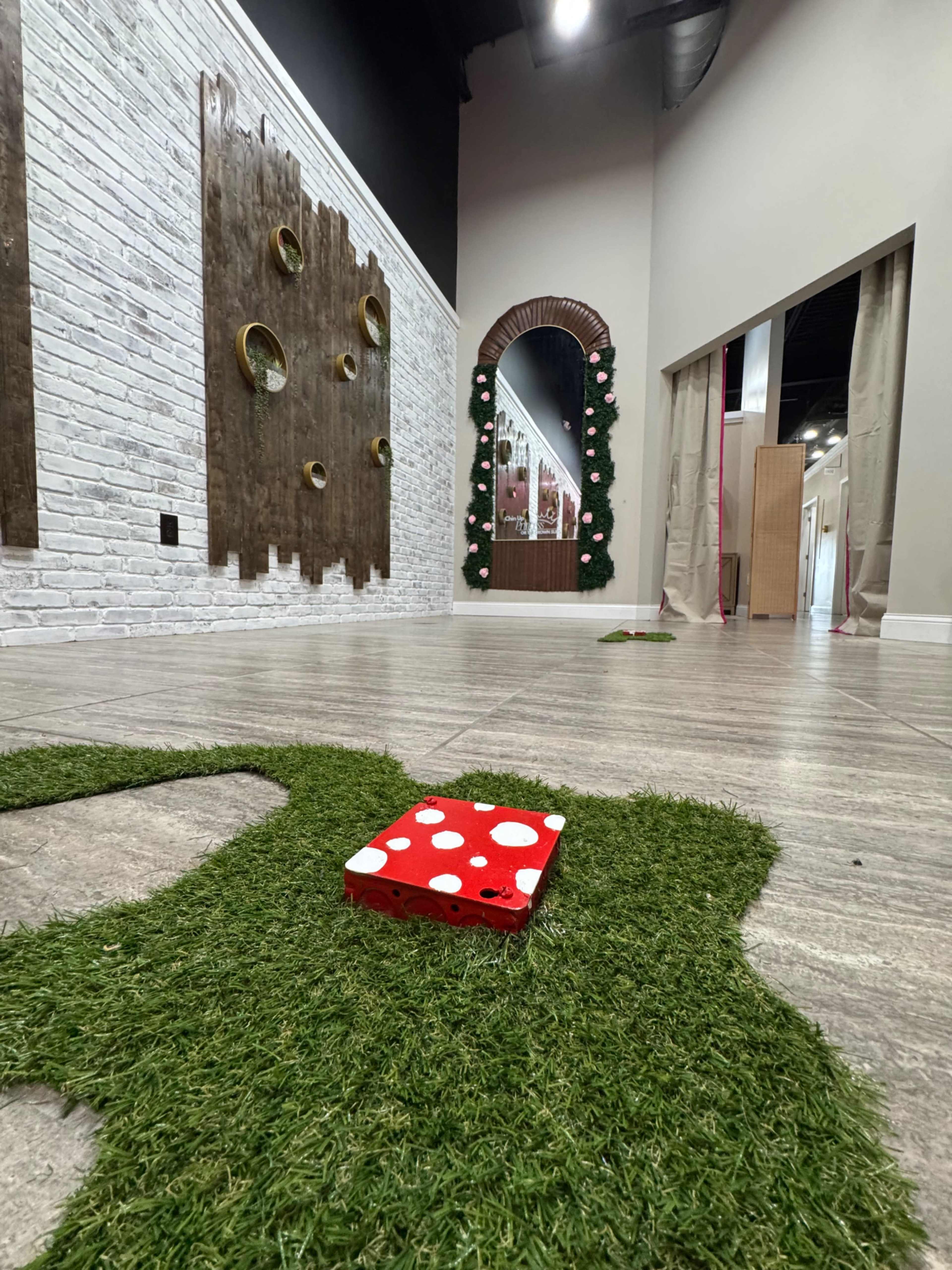 The image shows a modern interior space with a decorative wall featuring circular cutouts, a large mirror decorated with greenery, and a patch of artificial grass on the floor.