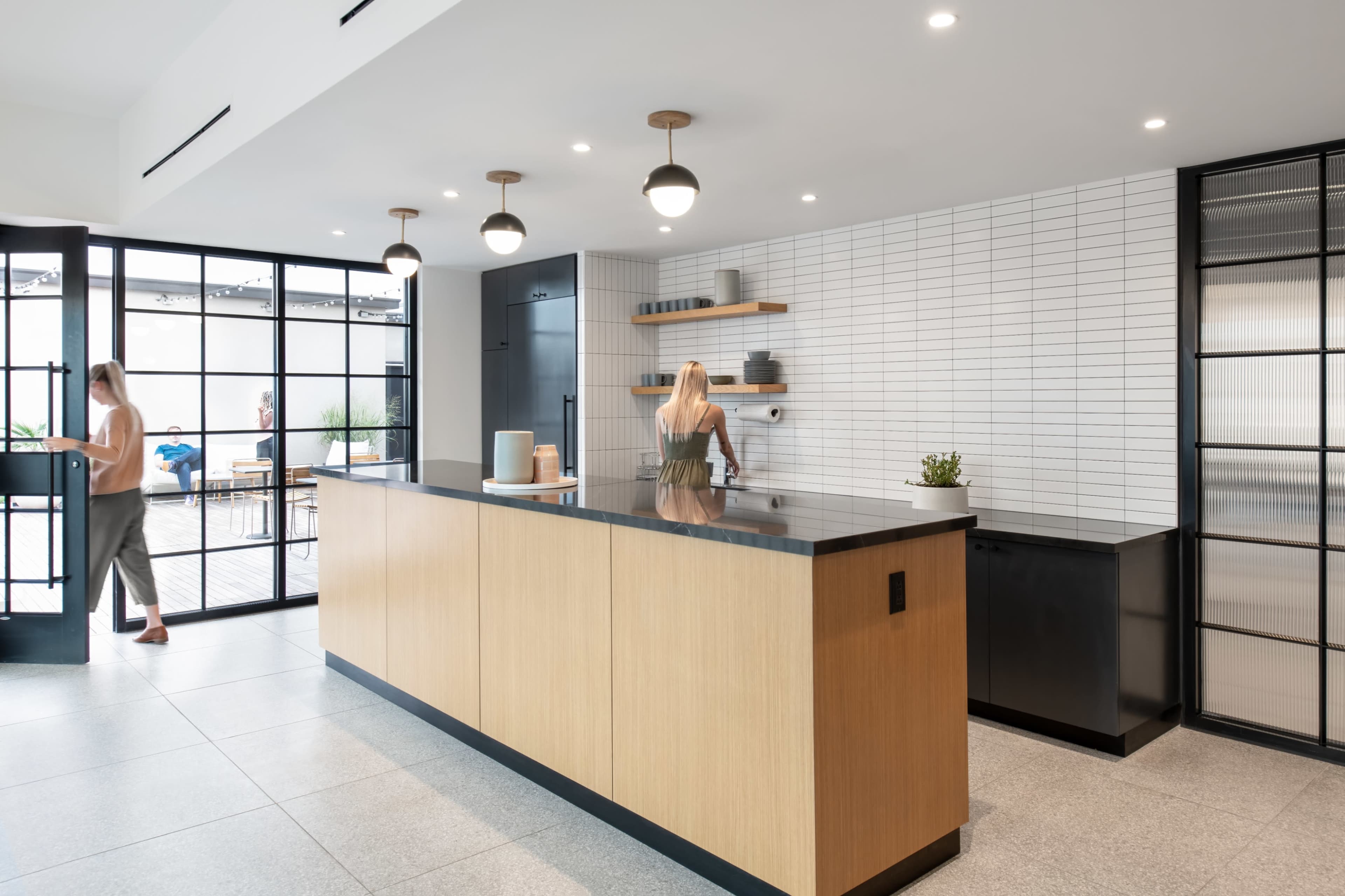 A modern kitchen features a large wooden island with a black countertop and shelves, surrounded by bright lighting and large glass doors leading to an outdoor area.