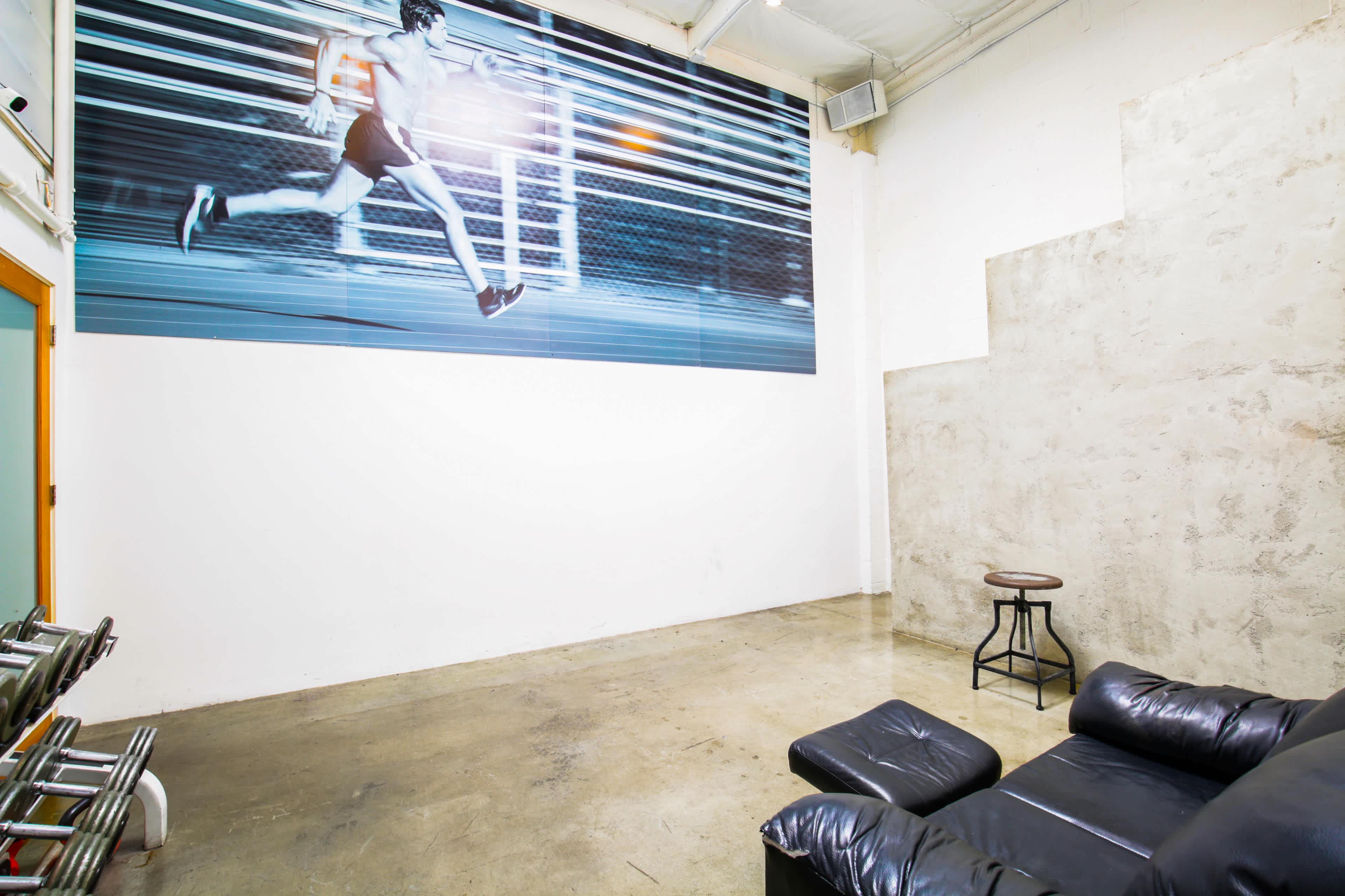 A spacious room with a large mural of a runner on one wall, a black leather couch, a stool, and exposed concrete surfaces.