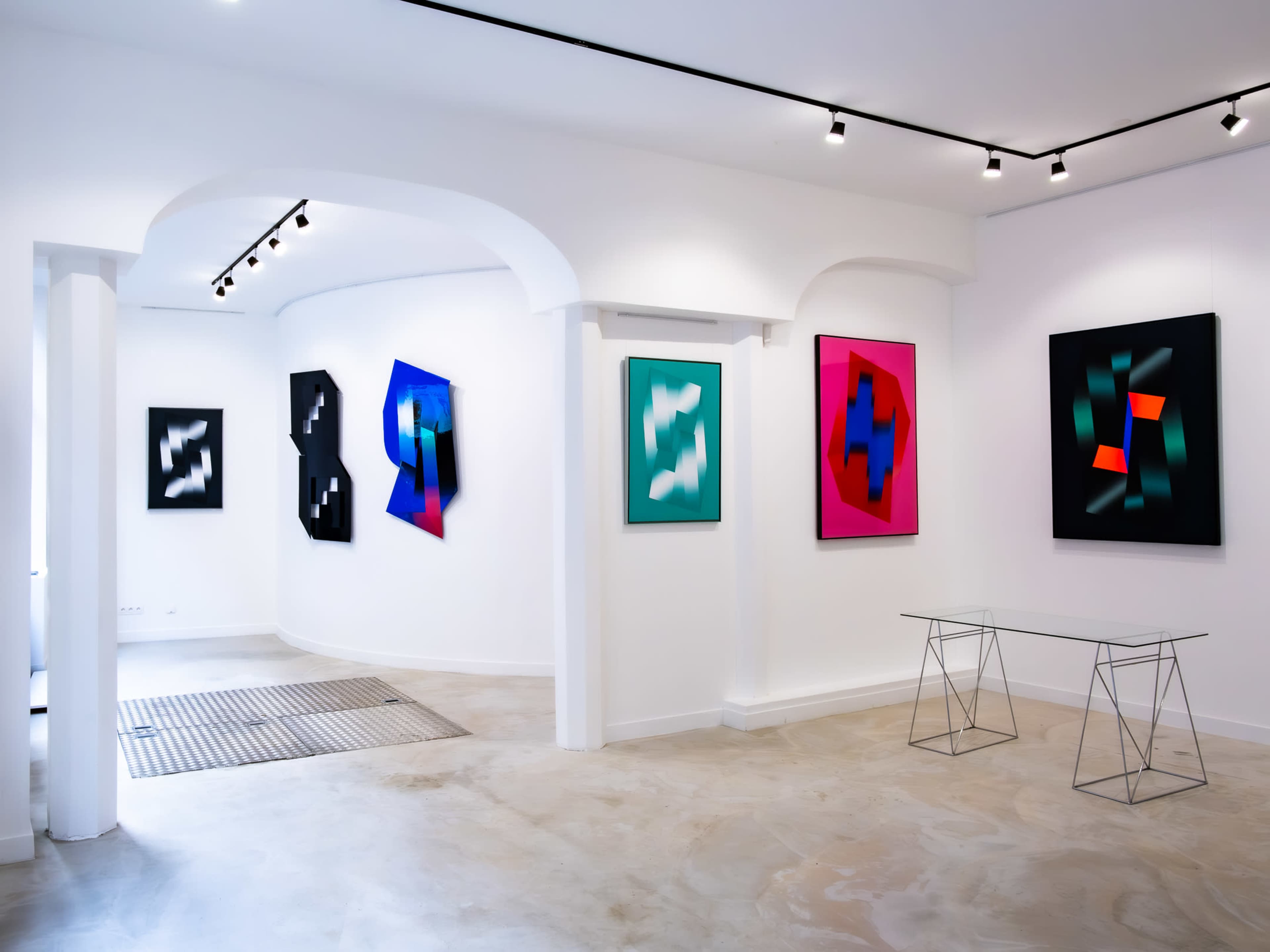 A modern art gallery with several colorful abstract paintings displayed on the white walls, and a minimalist glass table positioned in the center.