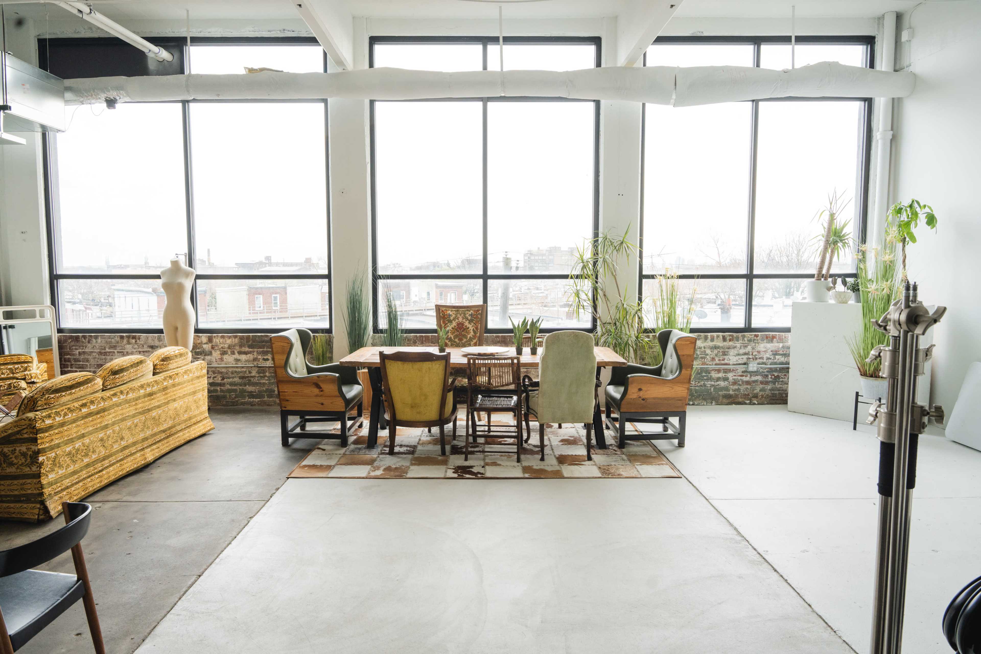 A spacious, well-lit room with large windows, featuring a dining table and chairs, a sofa, and several potted plants.