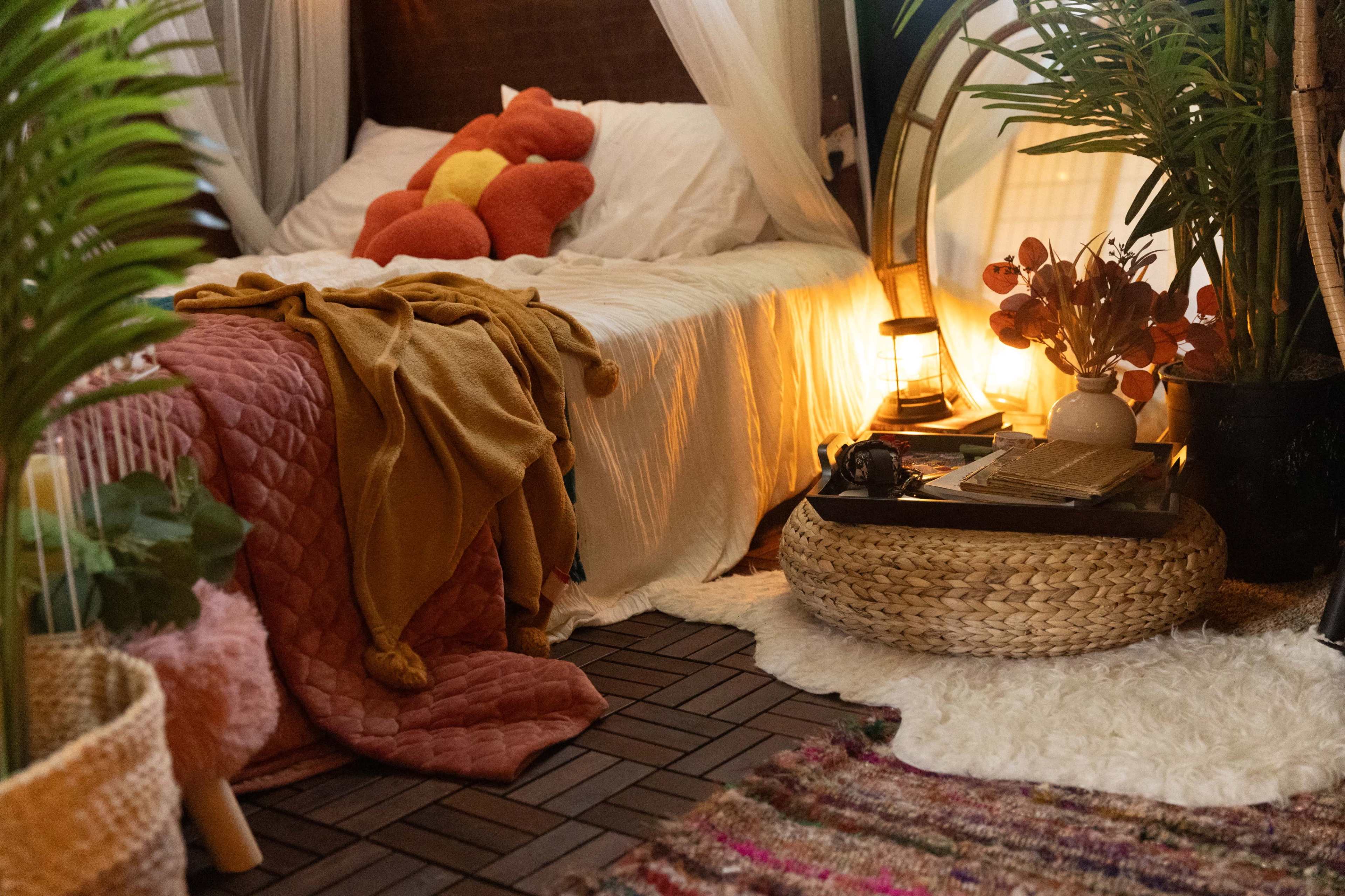 A cozy bedroom features a neatly made bed with colorful textiles, a lantern, and various plants arranged around a woven coffee table.