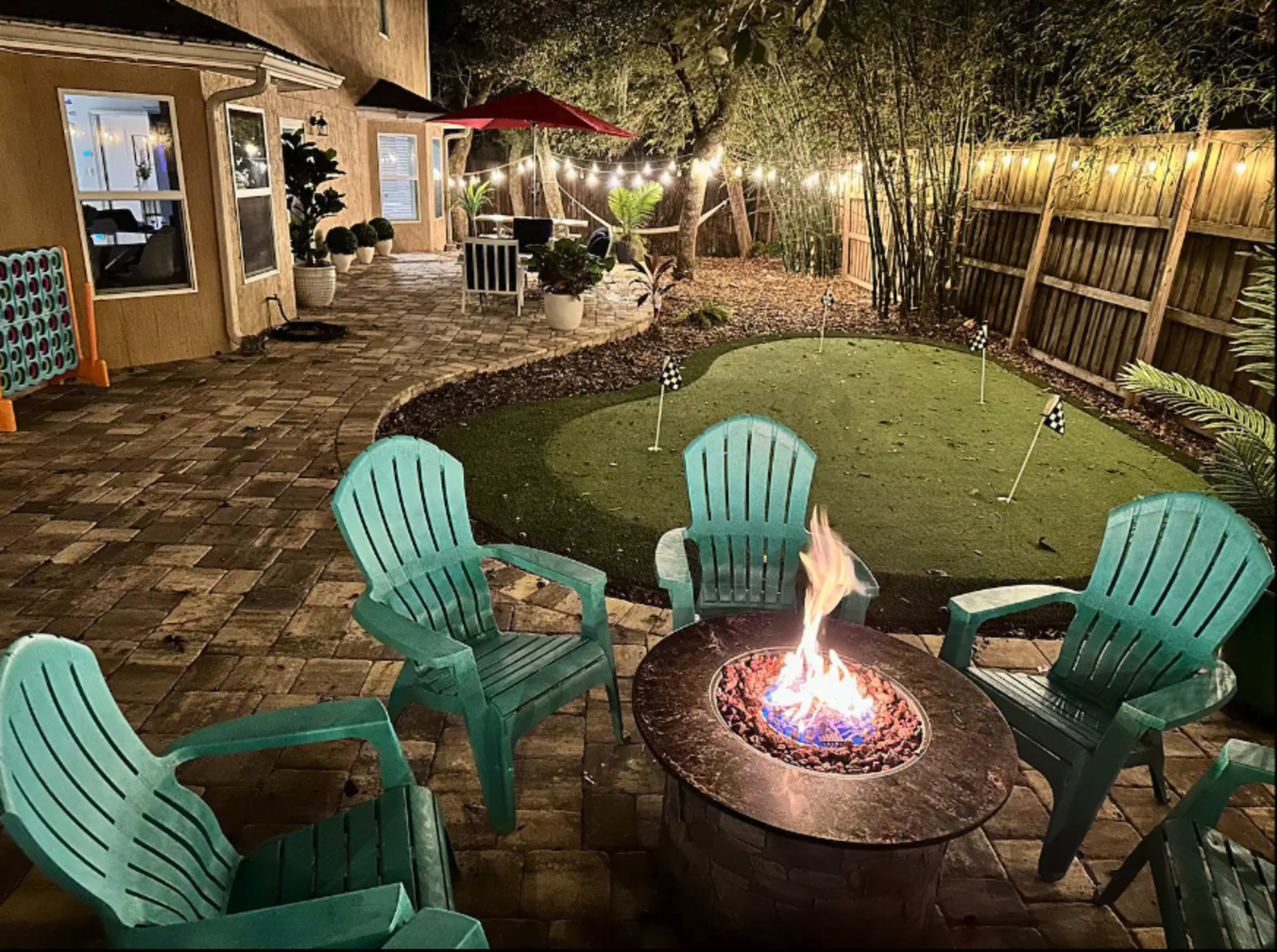 A stone fire pit surrounded by turquoise Adirondack chairs is located in a landscaped backyard with a putting green and string lights overhead.