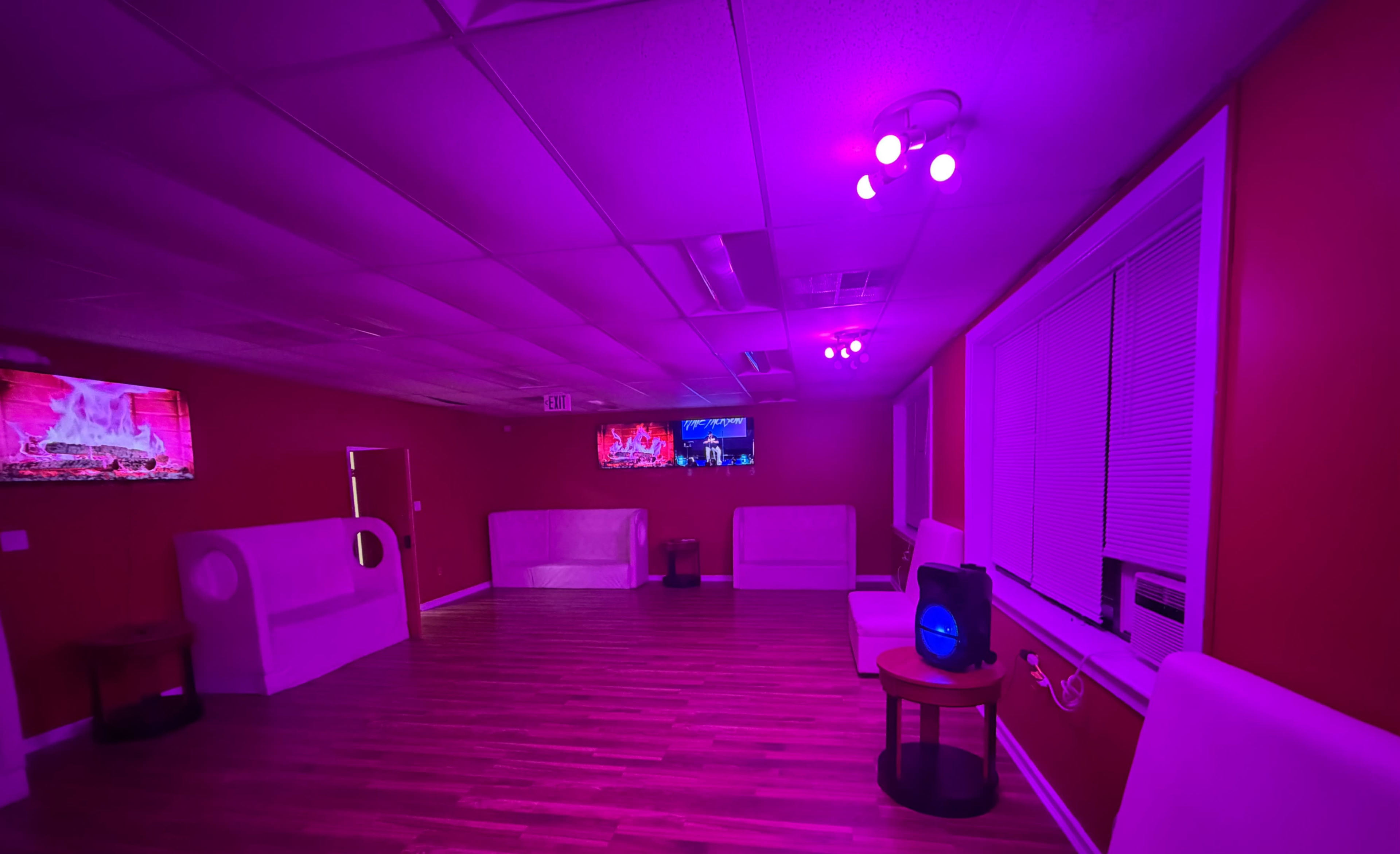 The image shows a dimly lit room with pink lighting, featuring white furniture, wooden flooring, and a television mounted on the wall.