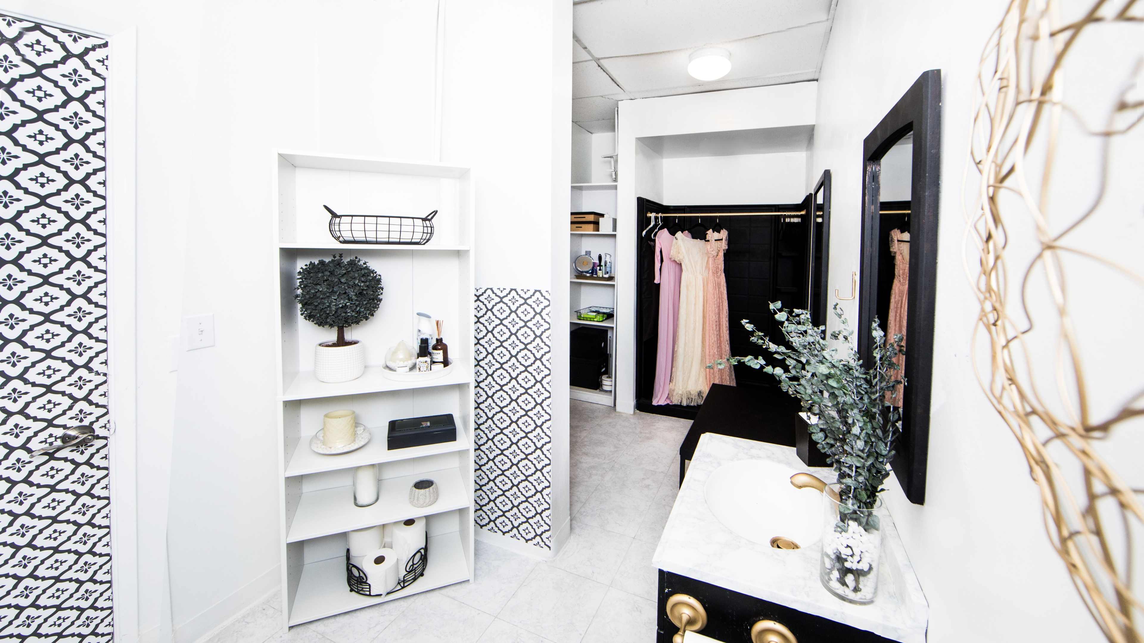 A modern bathroom with a white color scheme, featuring a decorative shelving unit, a vanity with a sink, and a closet area displaying hanging garments.