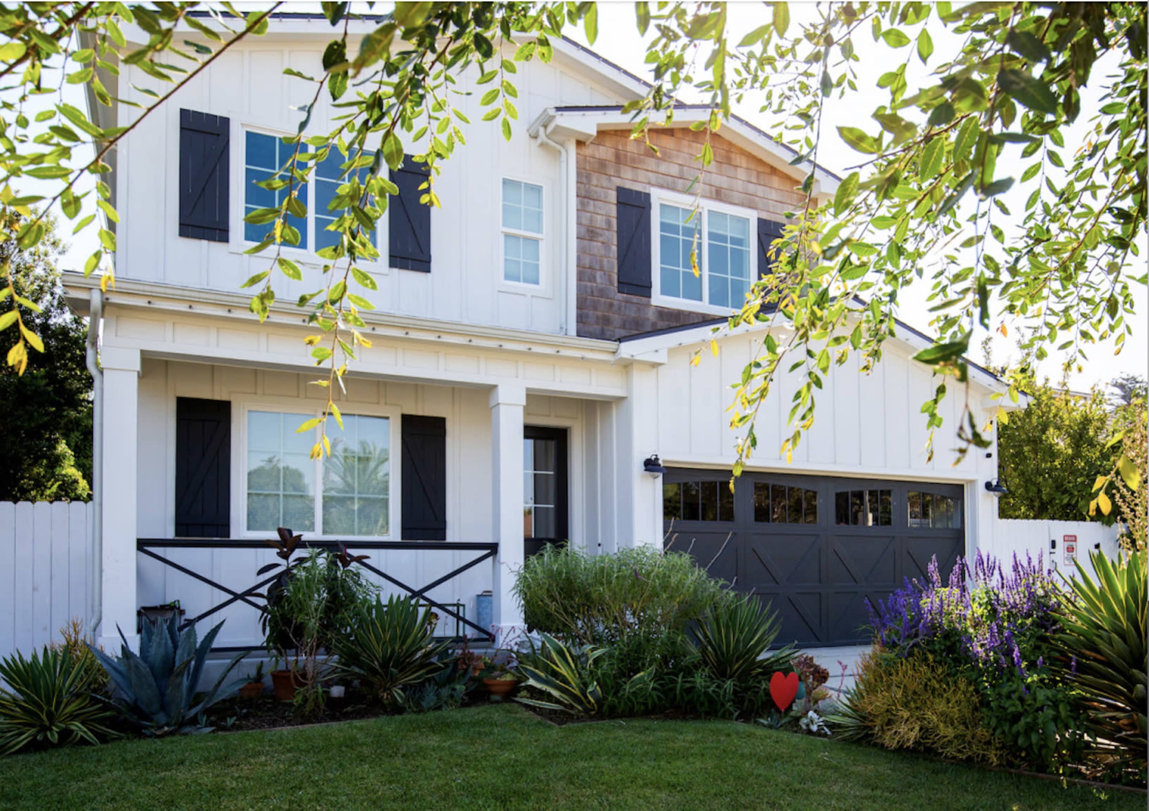 A two-story white house with black shutters and a brown shingle accent is surrounded by a well-maintained garden featuring various plants and a decorative heart.