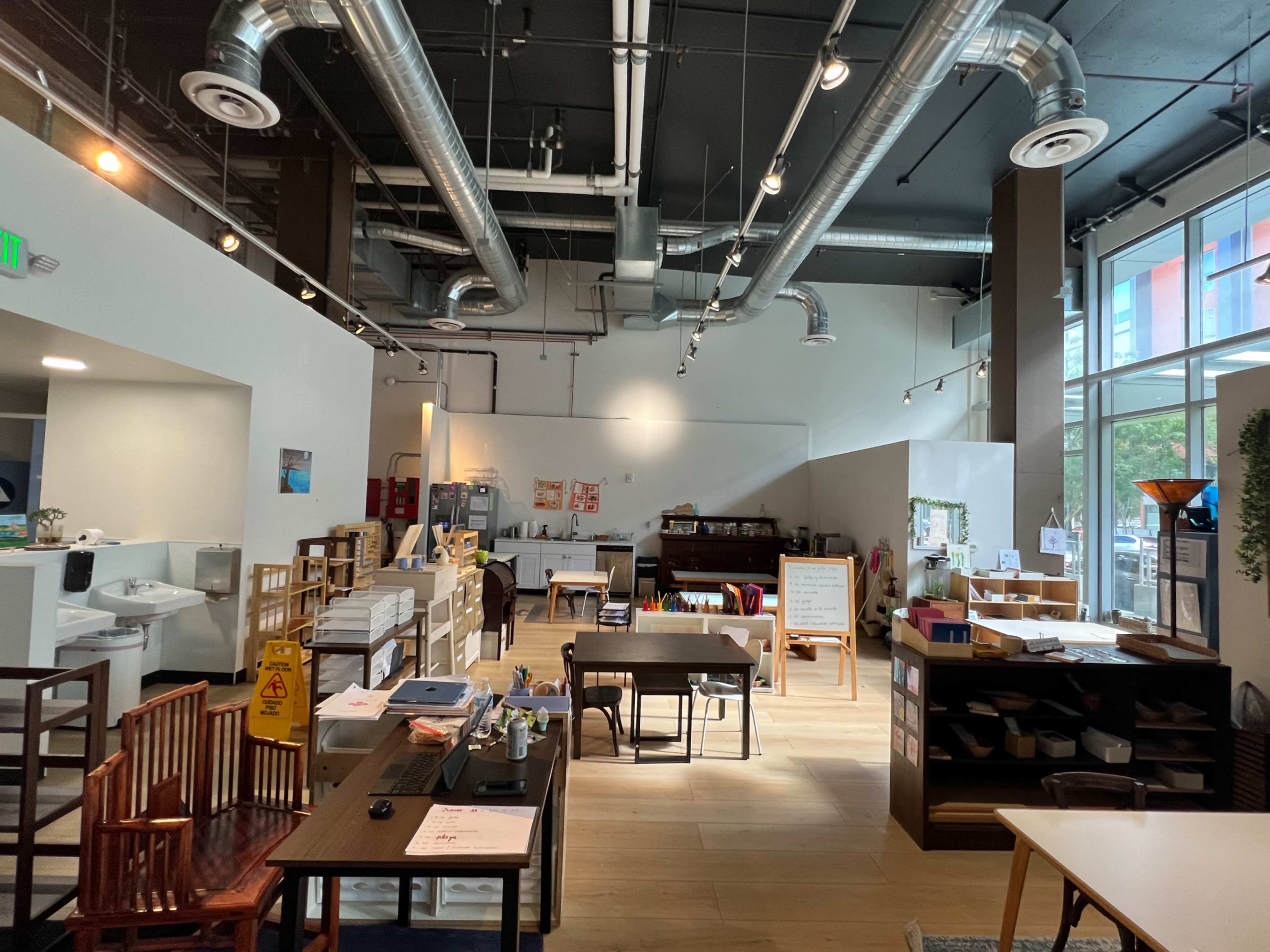 The image depicts a spacious, modern workspace with wooden tables, chairs, and shelves, illuminated by natural light from large windows.