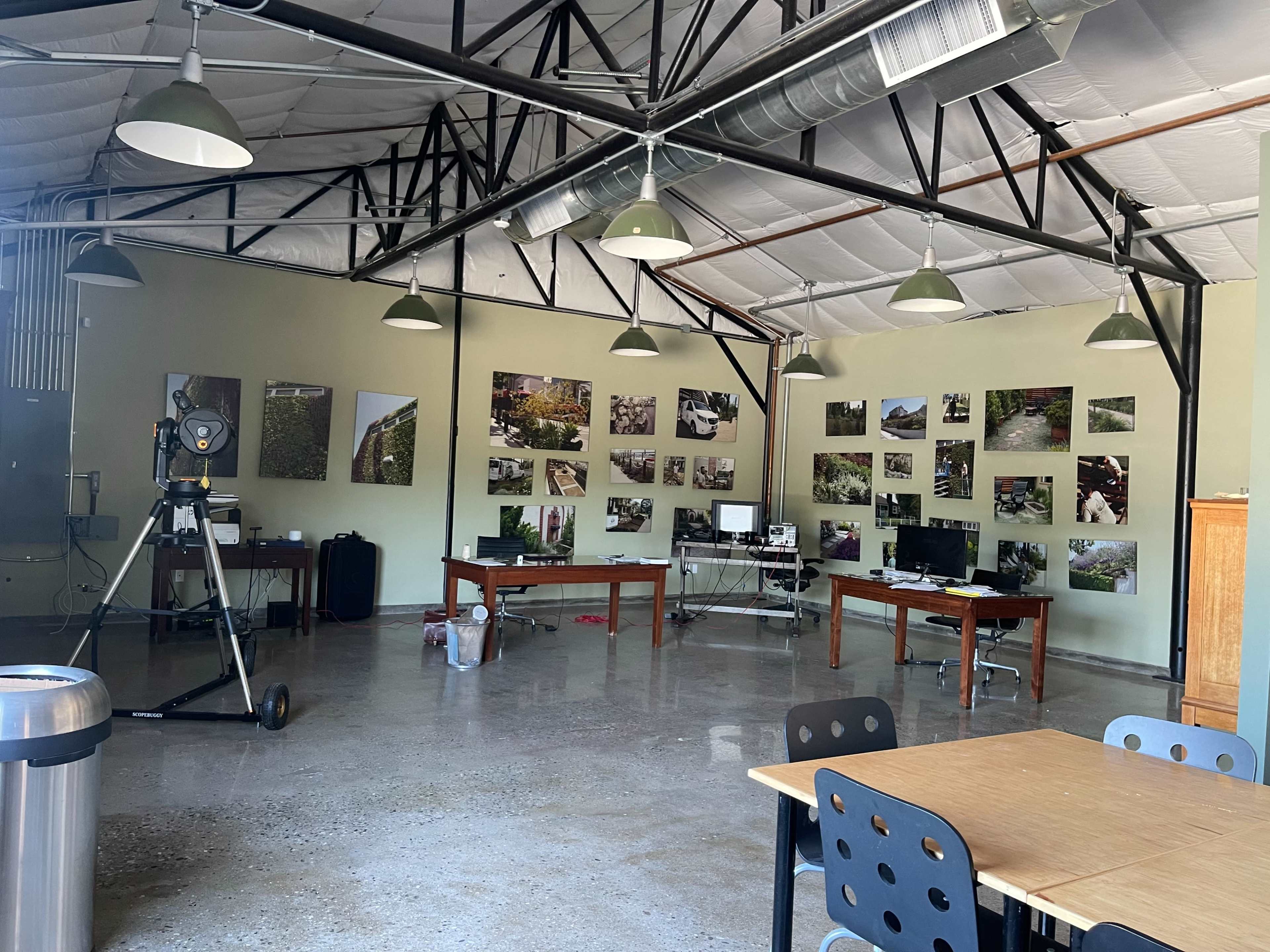 The image shows a spacious room with high ceilings, featuring several desks with computers and a large camera setup, surrounded by photographs displayed on the walls.
