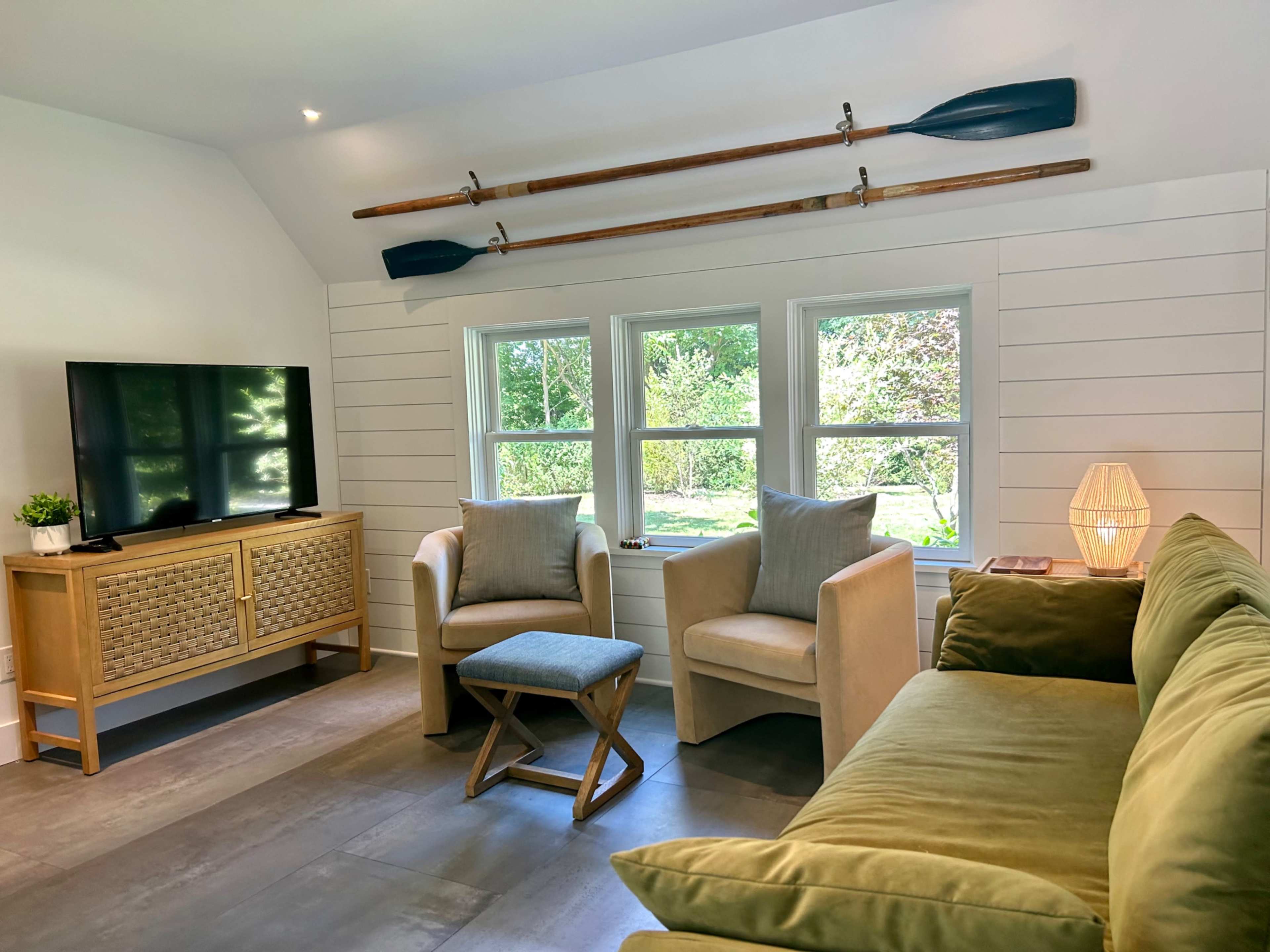 A cozy living room features a television, two armchairs, a small stool, and a green sofa, with decorative paddles mounted above the windows.