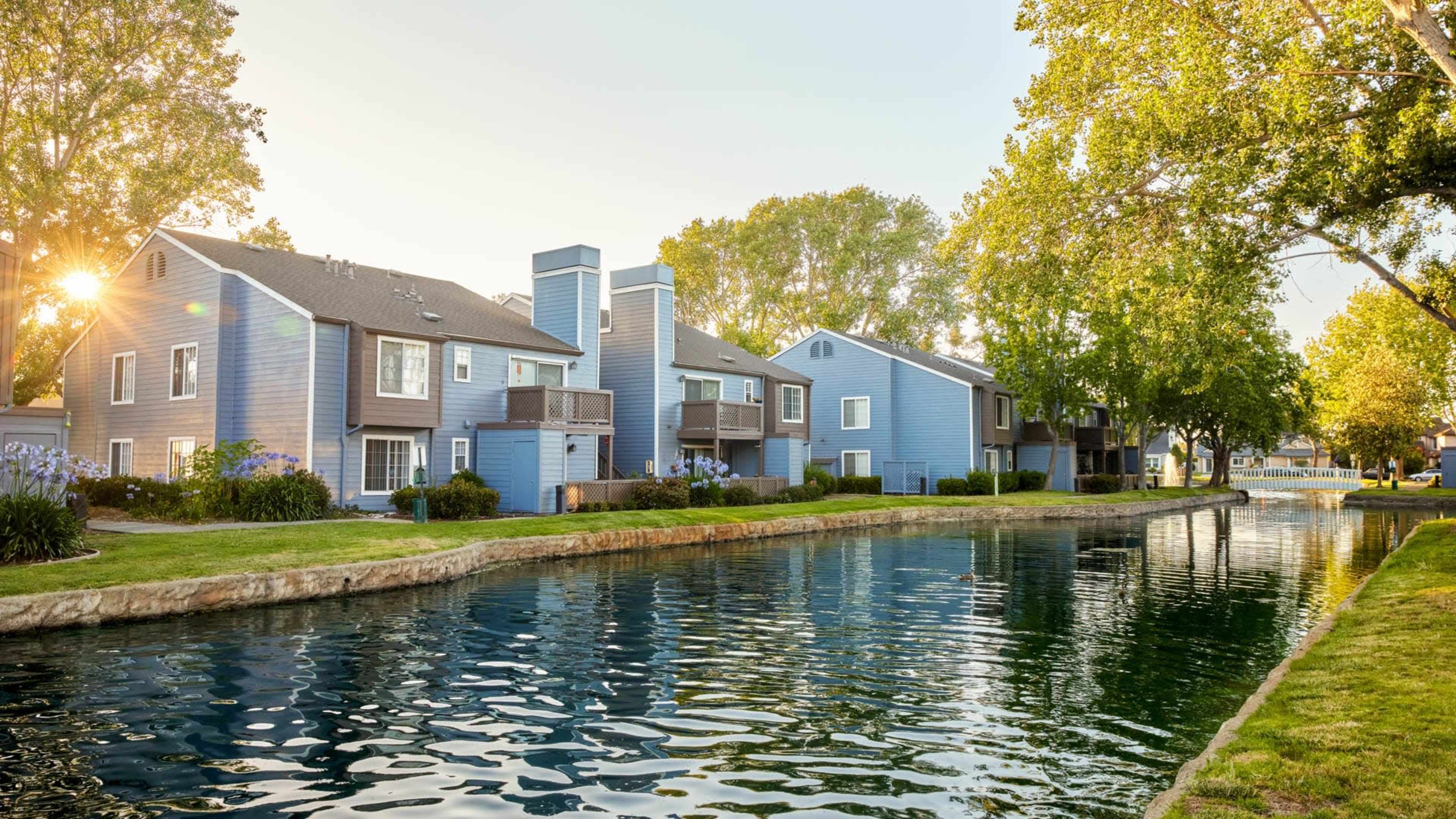 A row of blue residential buildings alongside a calm, reflective waterway surrounded by greenery.