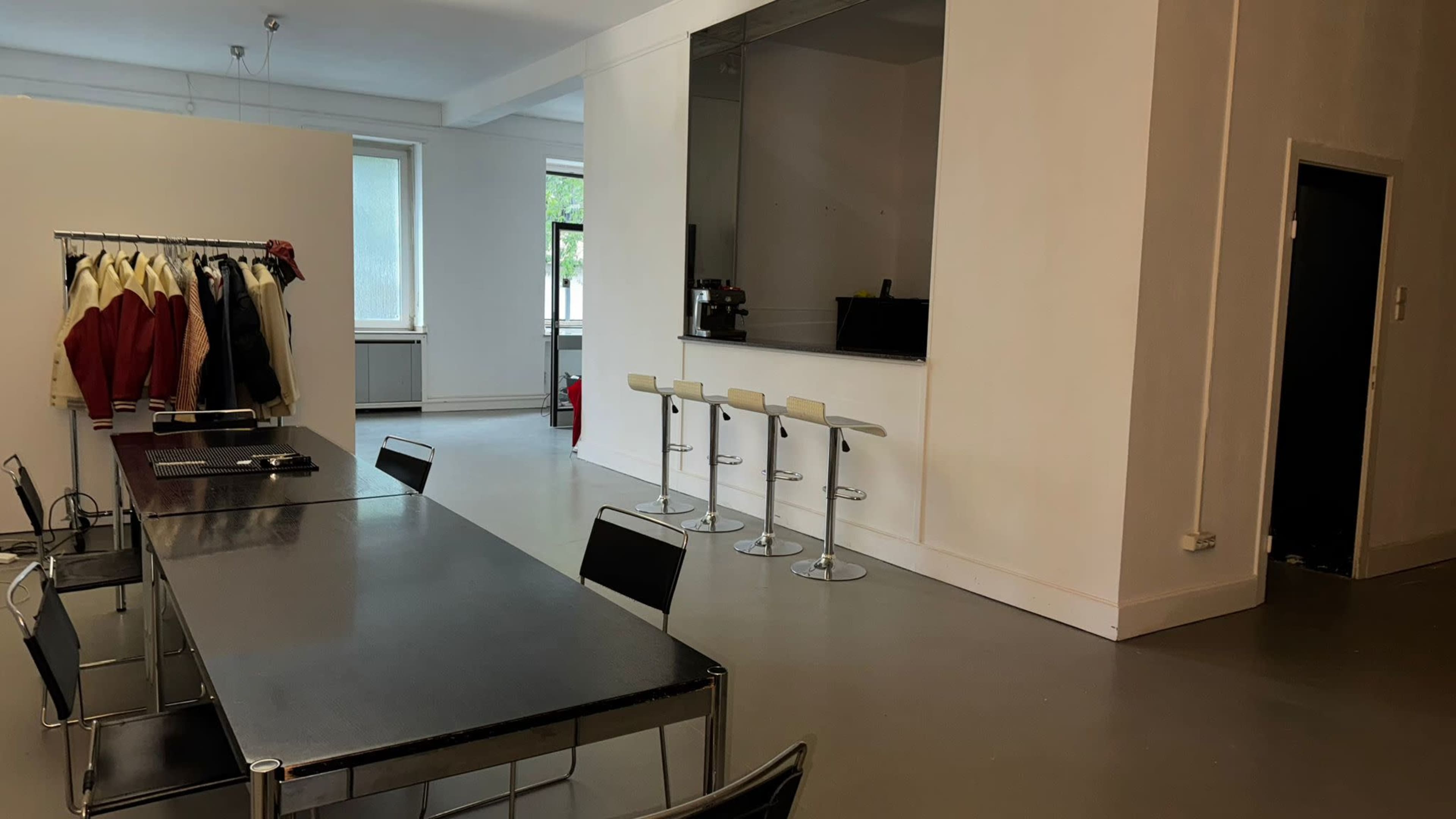 The image shows a spacious room with a long black dining table, several high stools arranged at a counter, and a clothing rack displaying jackets.