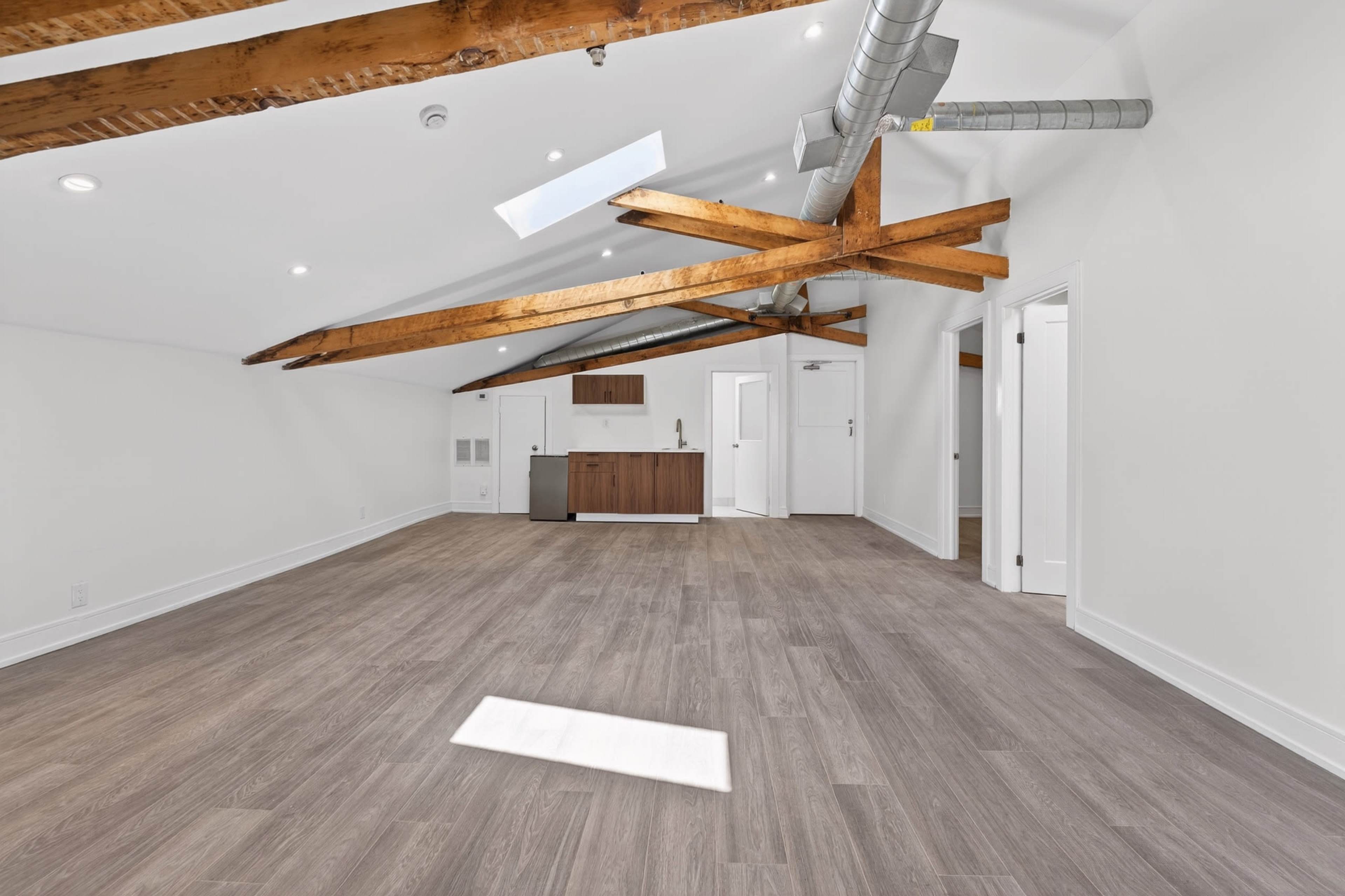 The image shows an empty interior room with wooden beams on the ceiling, light-colored walls, and a kitchen area in the background.