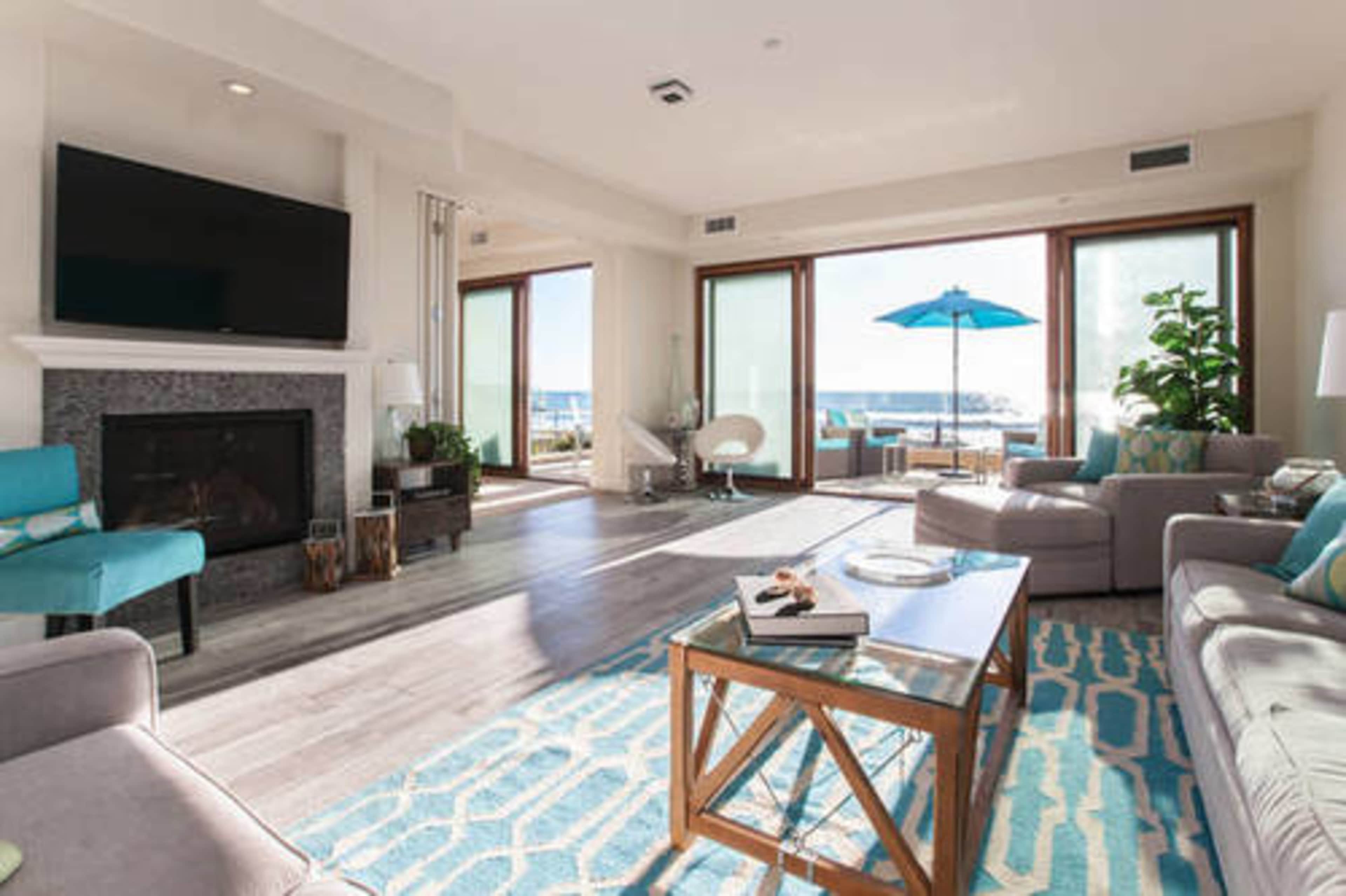 A bright, modern living room with large windows overlooking a patio featuring an umbrella, a comfortable seating area, and a fireplace.