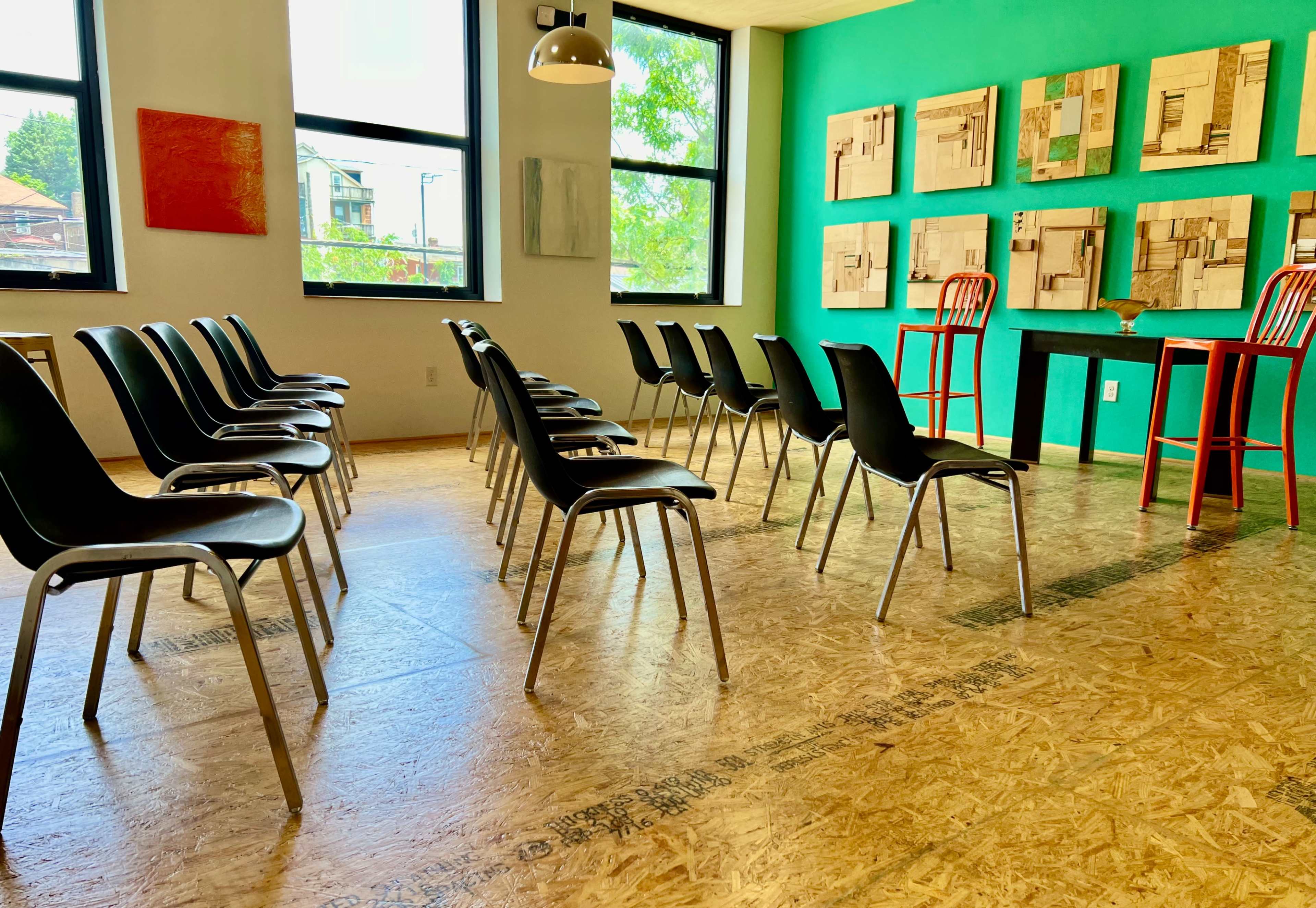A room with several black chairs arranged in rows facing a green wall adorned with framed artwork, and two red chairs positioned at a small table.