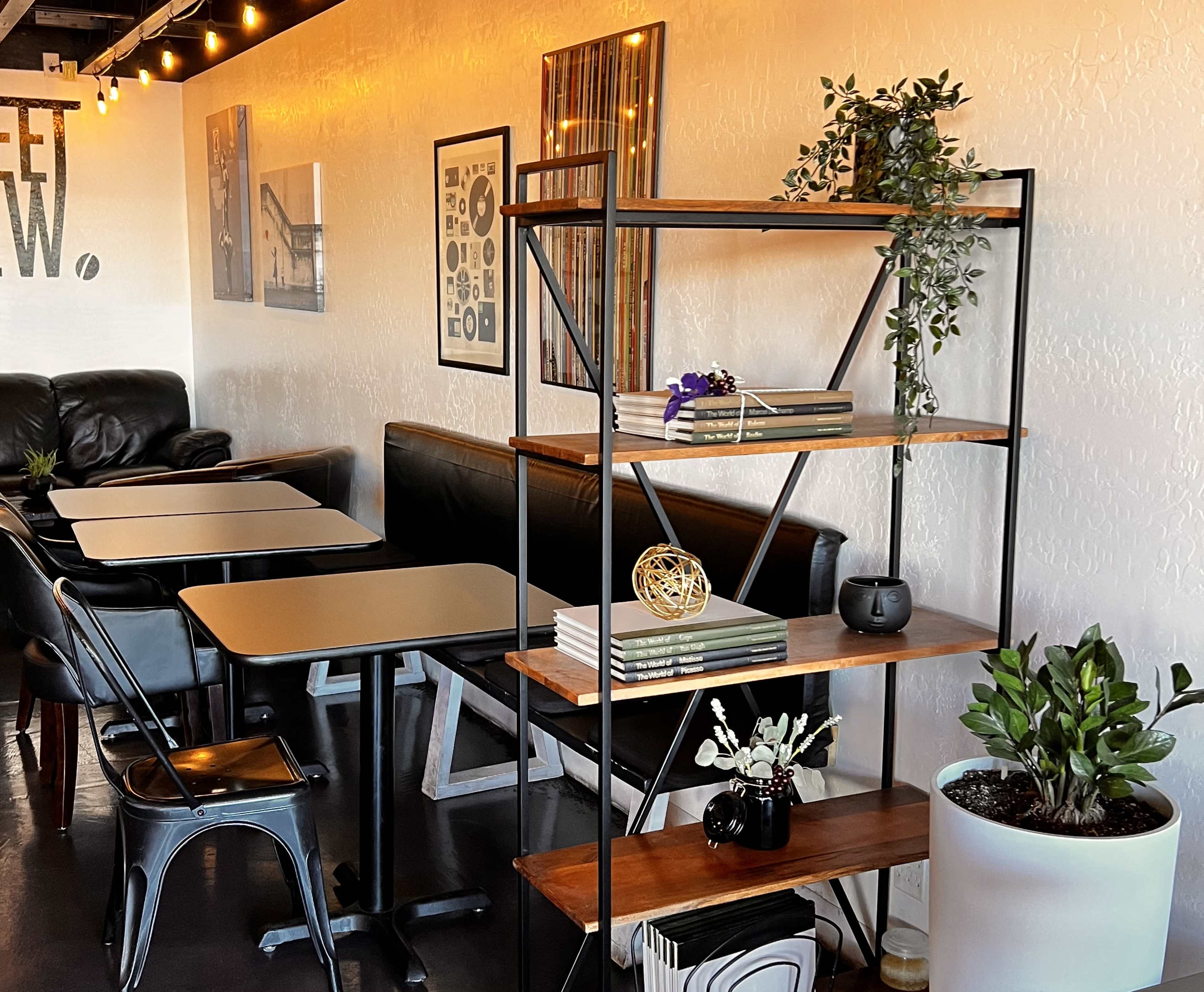 A metal and wood shelving unit stands next to a seating area with tables and chairs in a modern cafe.