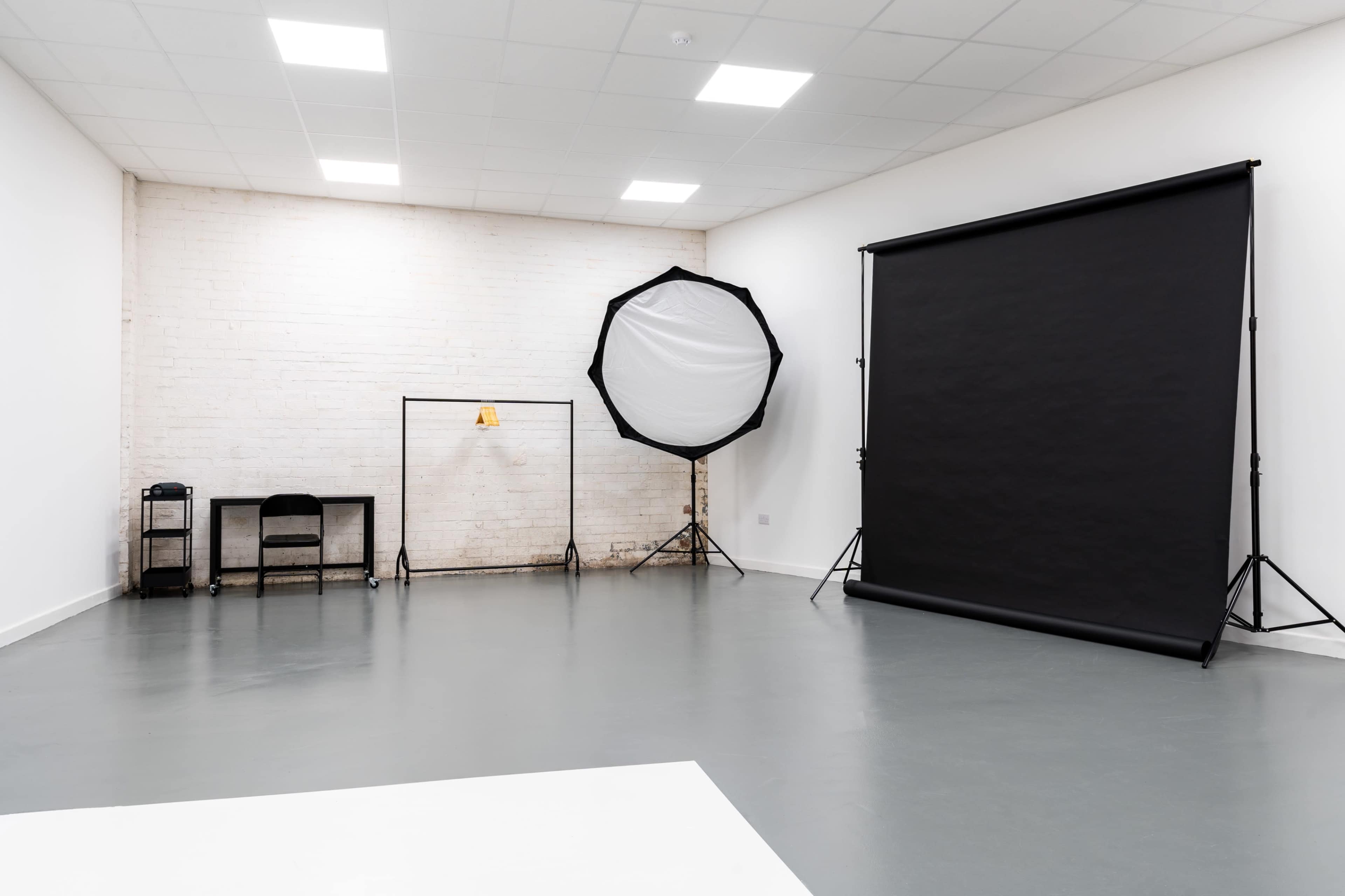 The image shows a photography studio featuring a black backdrop, a large softbox light, and minimal furniture against a plain wall.