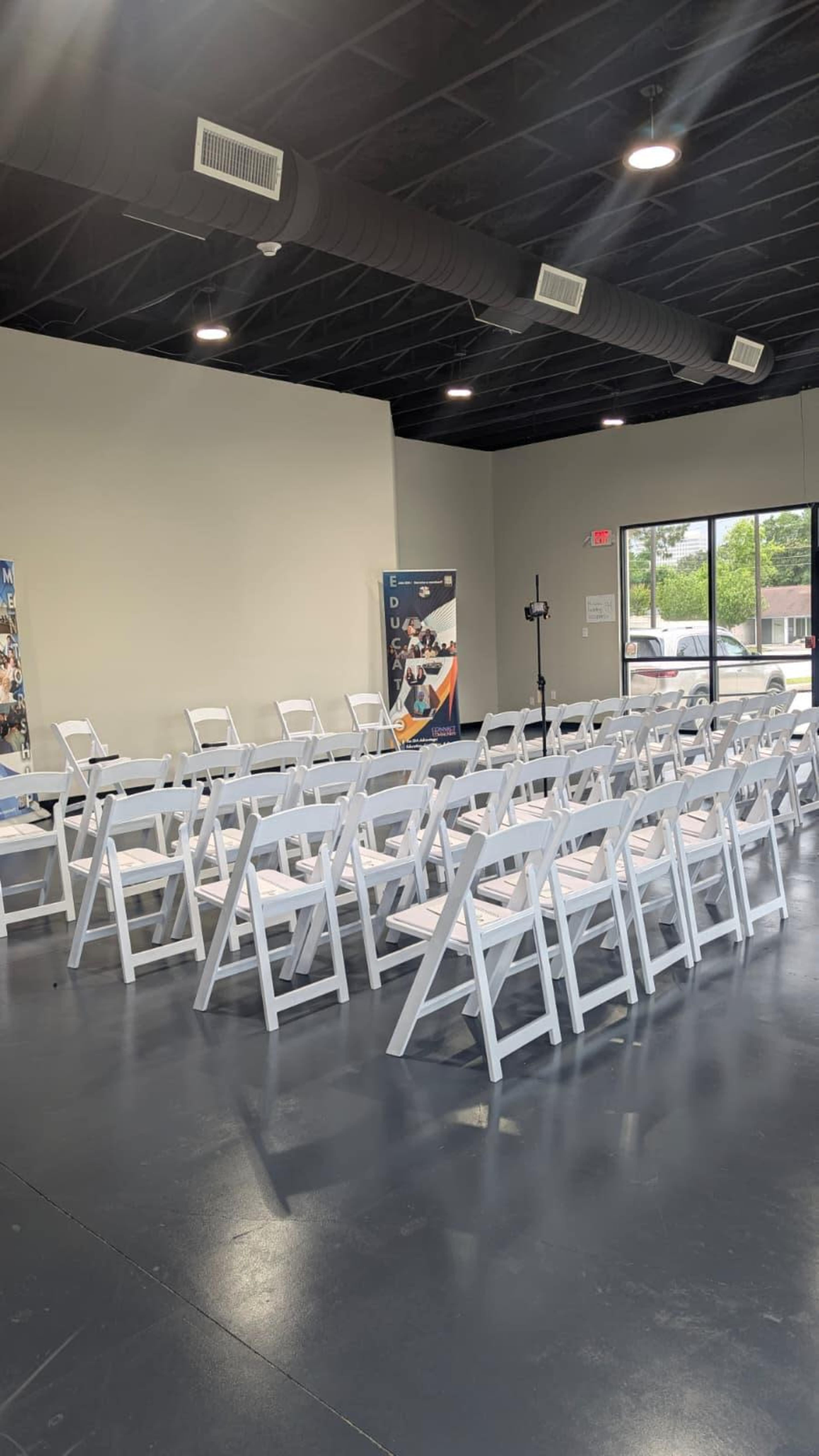 The image shows a spacious indoor area set up with rows of white folding chairs facing a front wall, with banners displayed along the sides.