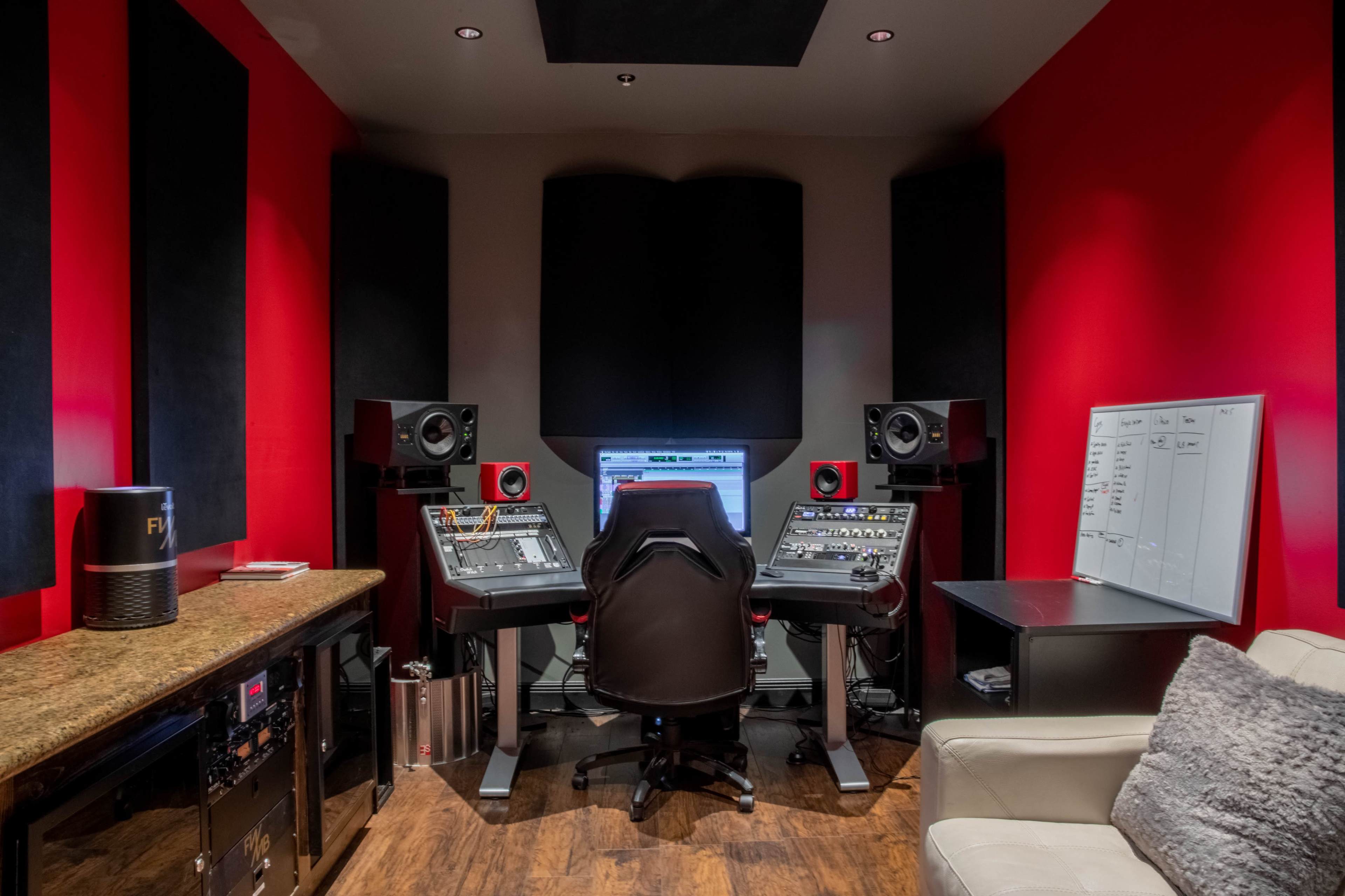 A music production studio features two control desks with equipment, large speakers, and a computer setup against red walls.