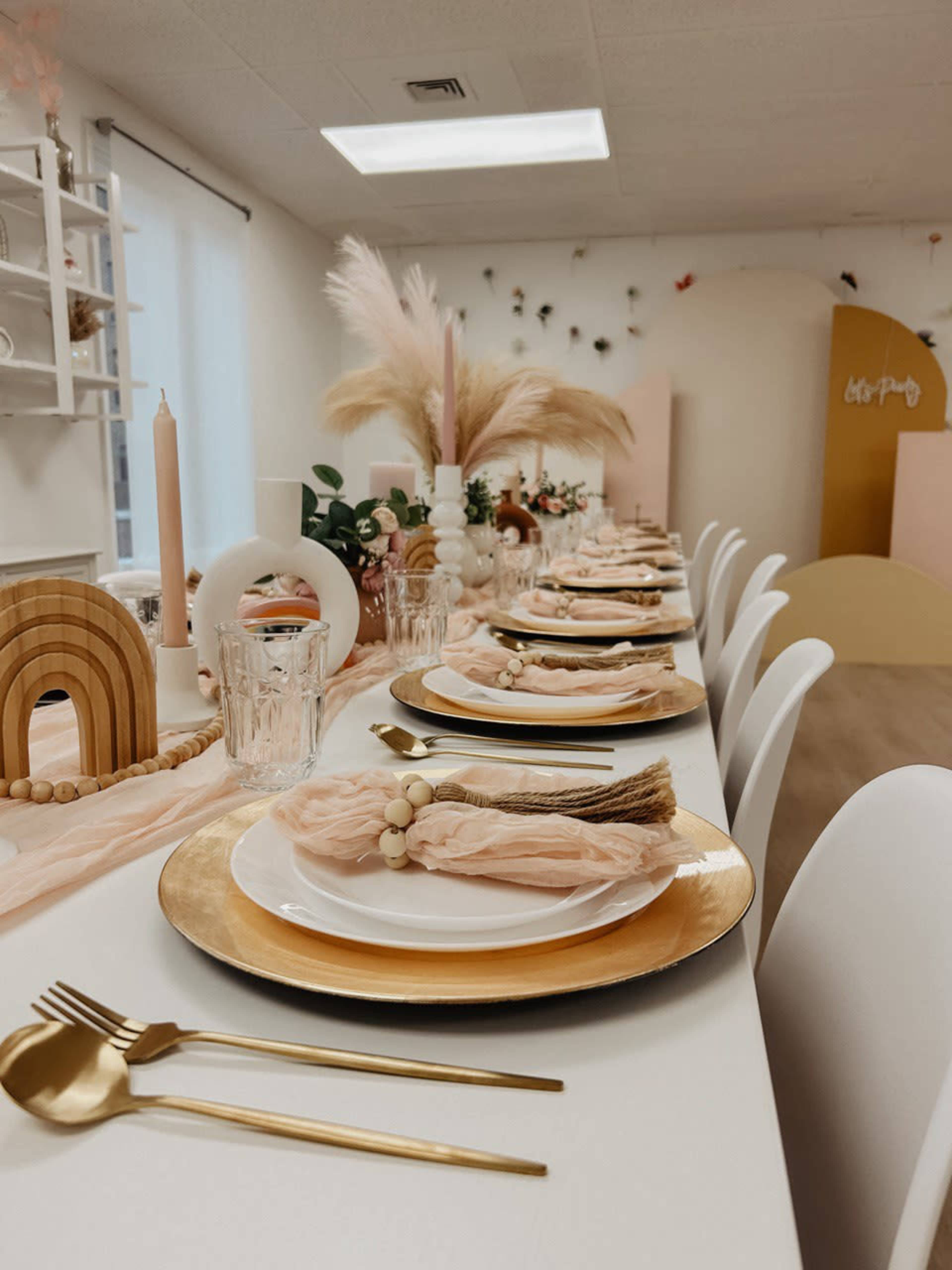 A long dining table is set with gold chargers, white plates, and blush napkins in an elegantly decorated room featuring pastel accents and decor.