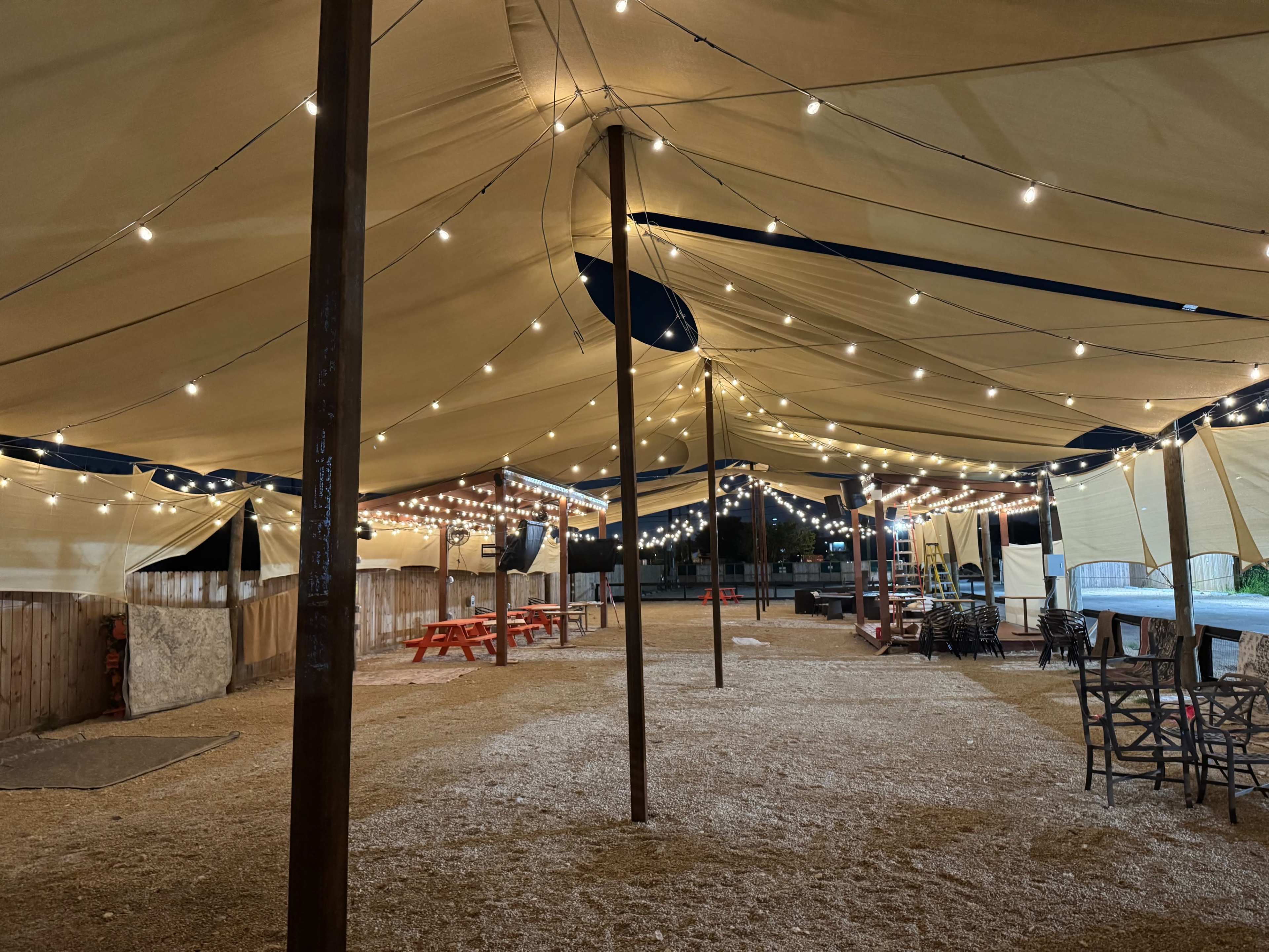 A spacious outdoor area is covered by a large tent with string lights illuminating picnic tables and chairs scattered across the gravel ground.