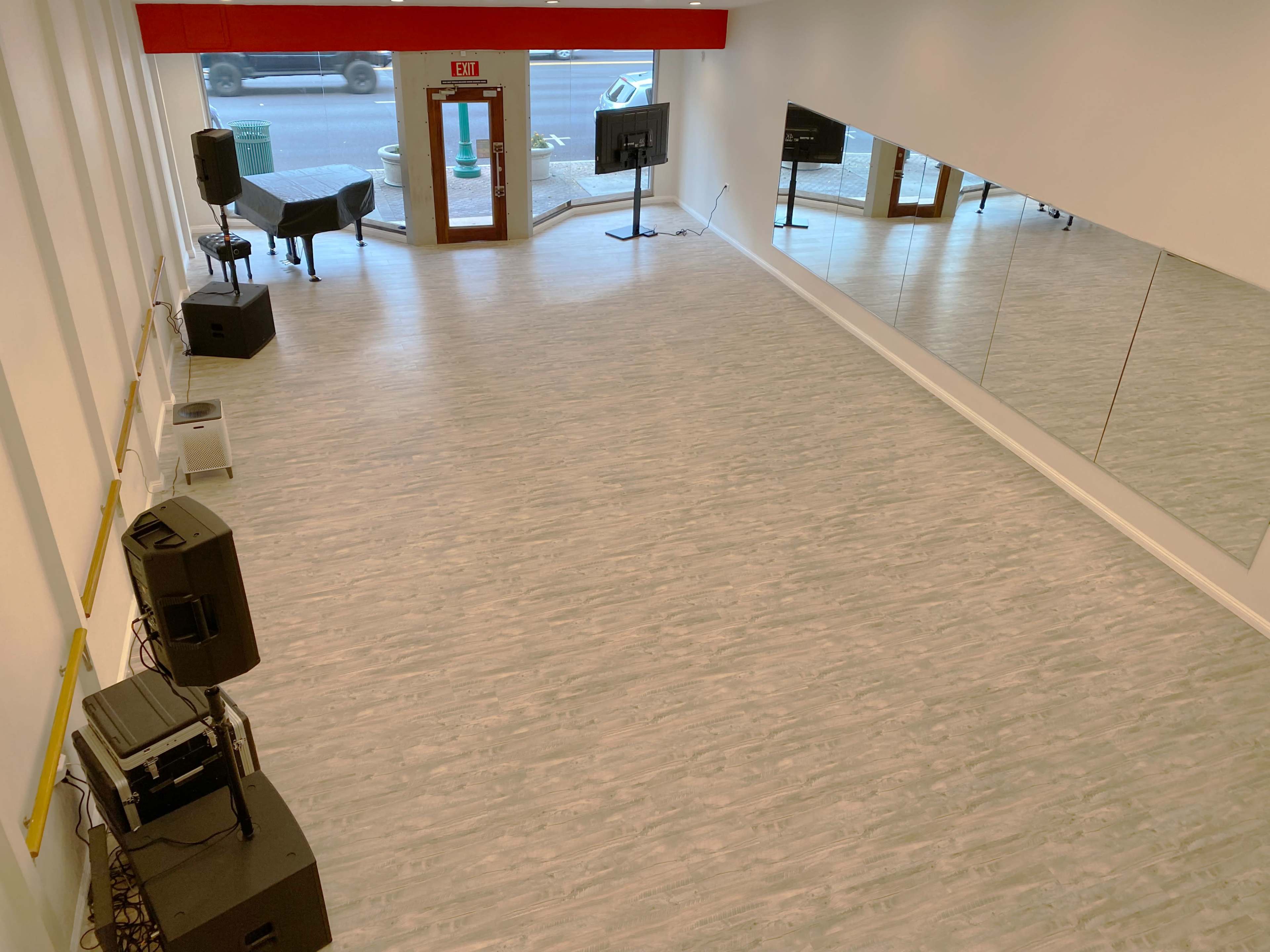 A spacious, empty room with light-colored flooring, large mirrors on one wall, a table and chair in the corner, and audio equipment set up at the front.