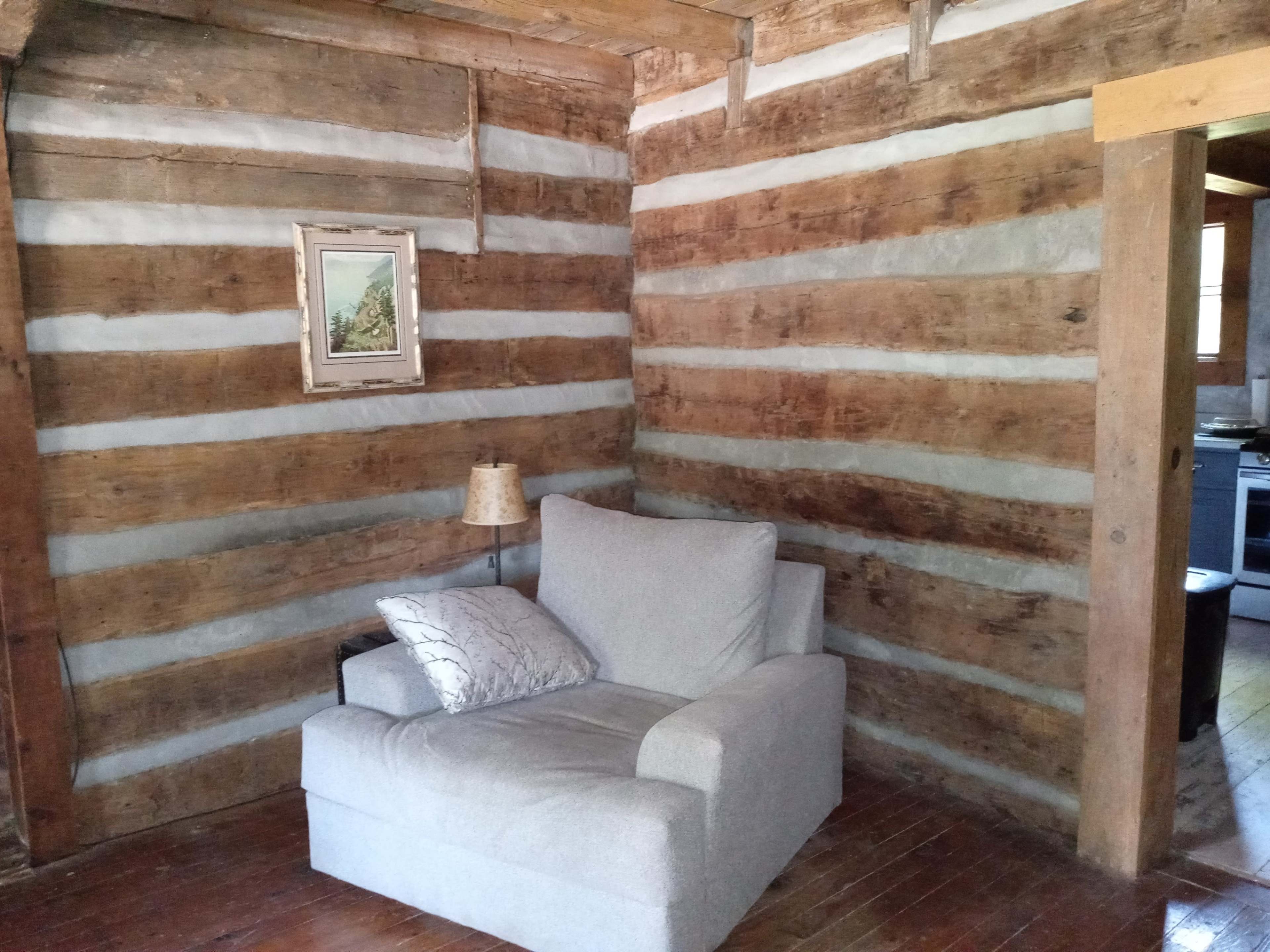 The image shows a cozy corner of a room with a gray armchair, a small lamp, a framed picture on the log walls, and a doorway leading to another space.