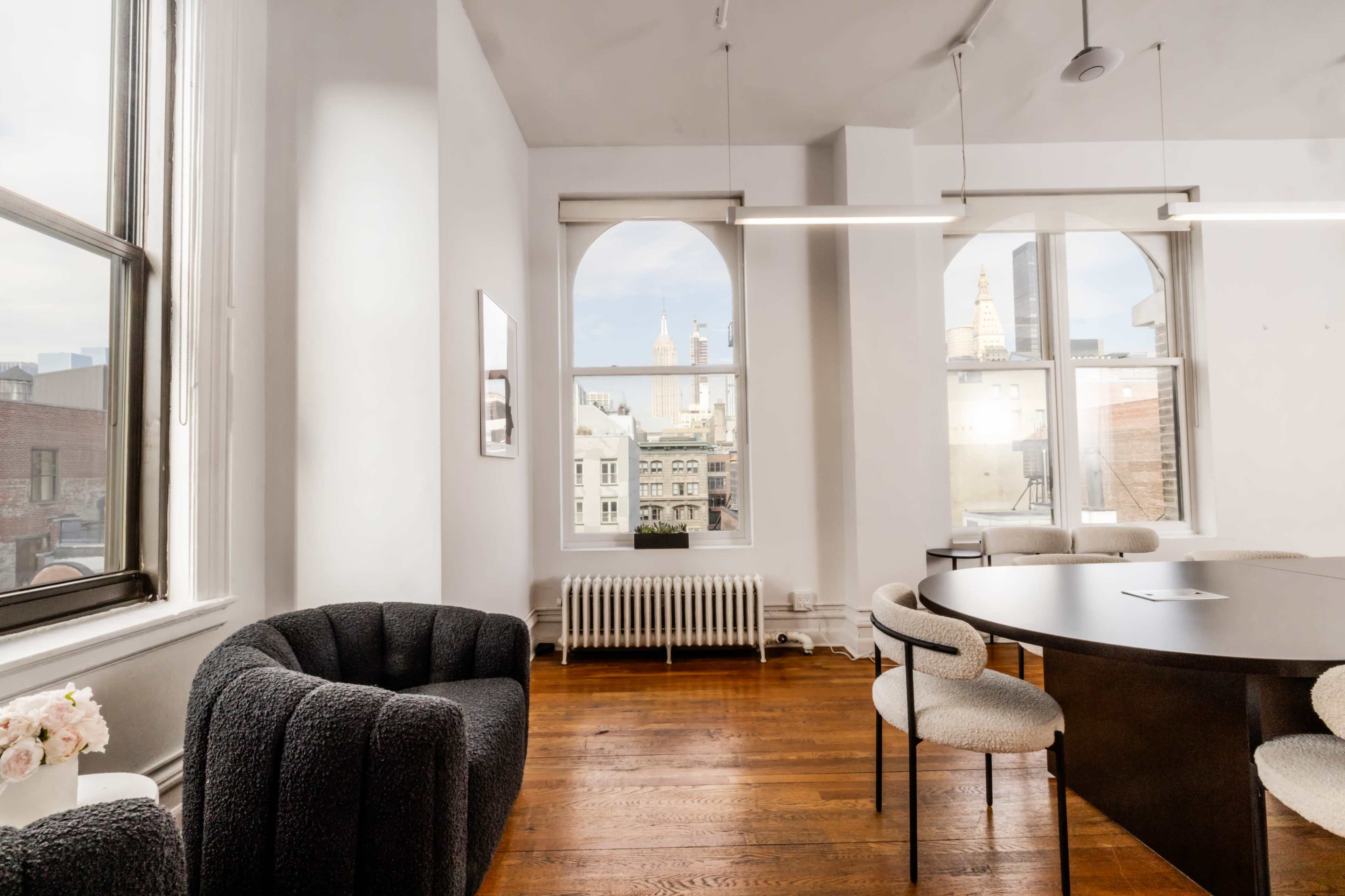 A well-lit modern office space features a round table with chairs, a cozy armchair, and large windows overlooking city buildings.