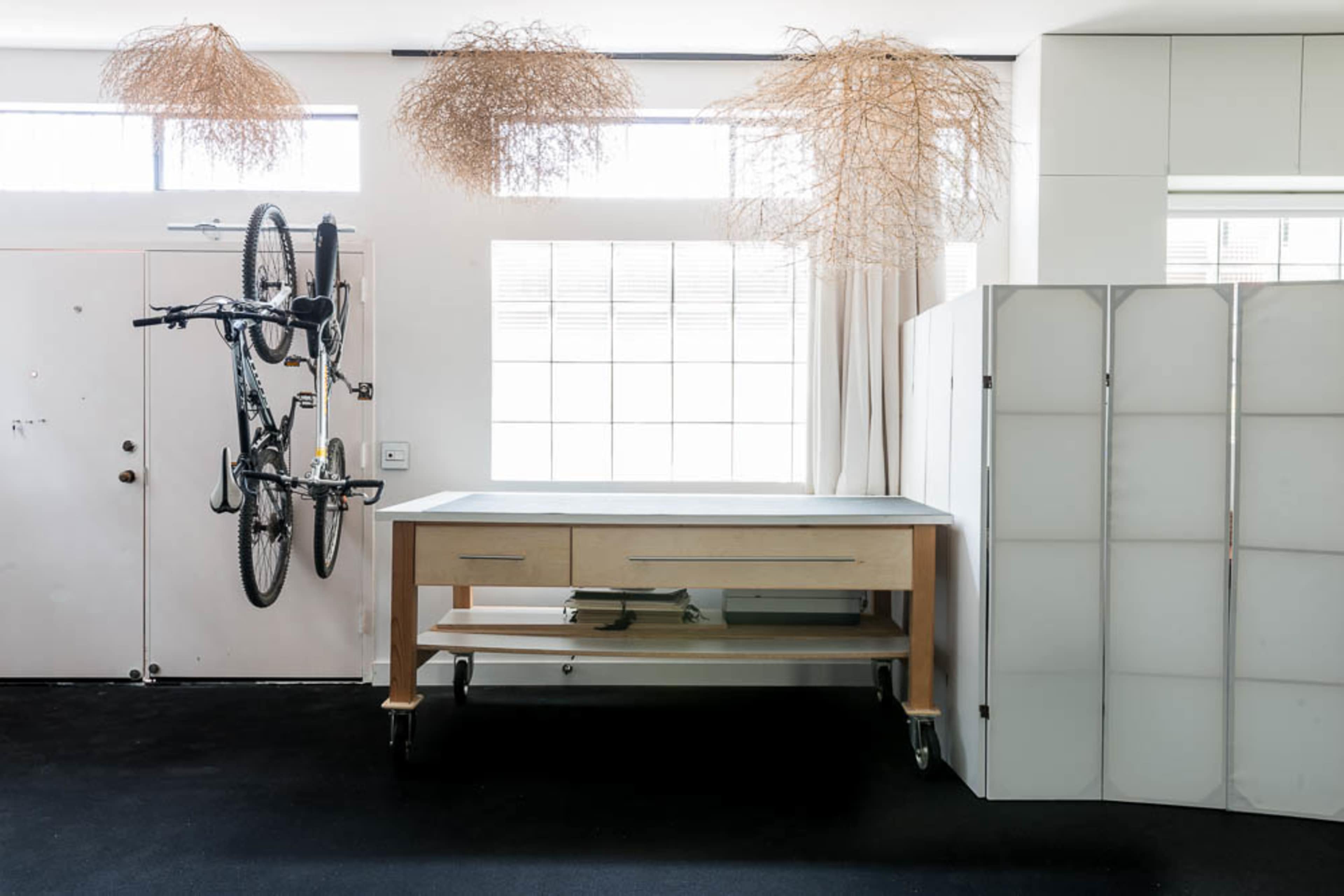 A workspace with a large wooden table on wheels, a bicycle hanging on the wall, and decorative dried grass arrangements overhead, positioned within a well-lit room.