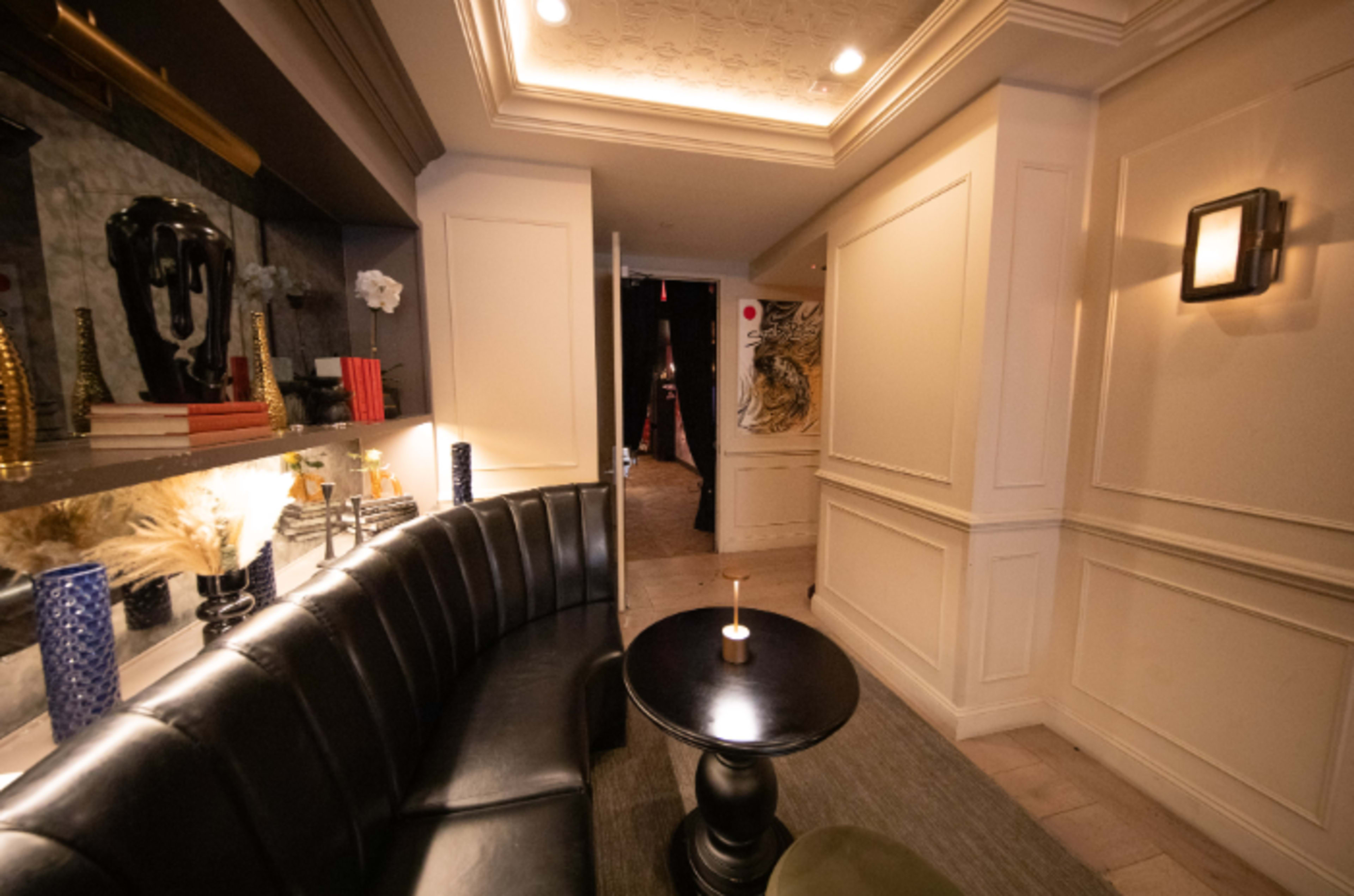 The image shows a stylish, semi-enclosed corridor with walls adorned in light colors, featuring a curved black leather bench, a small black table, and decorative elements like books and vases.