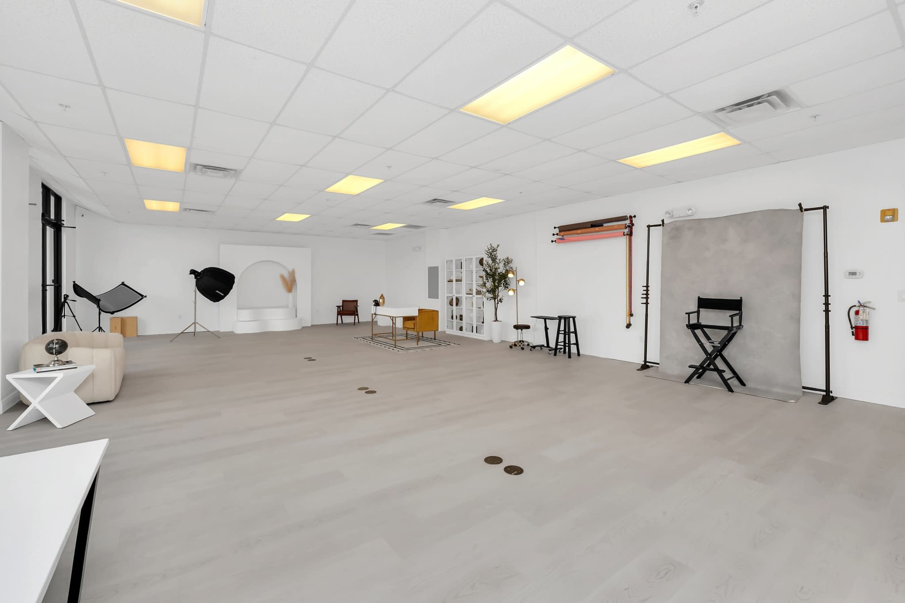The image shows a spacious, minimalist photography studio featuring a light-colored wooden floor, various lighting equipment, and a neutral backdrop area.