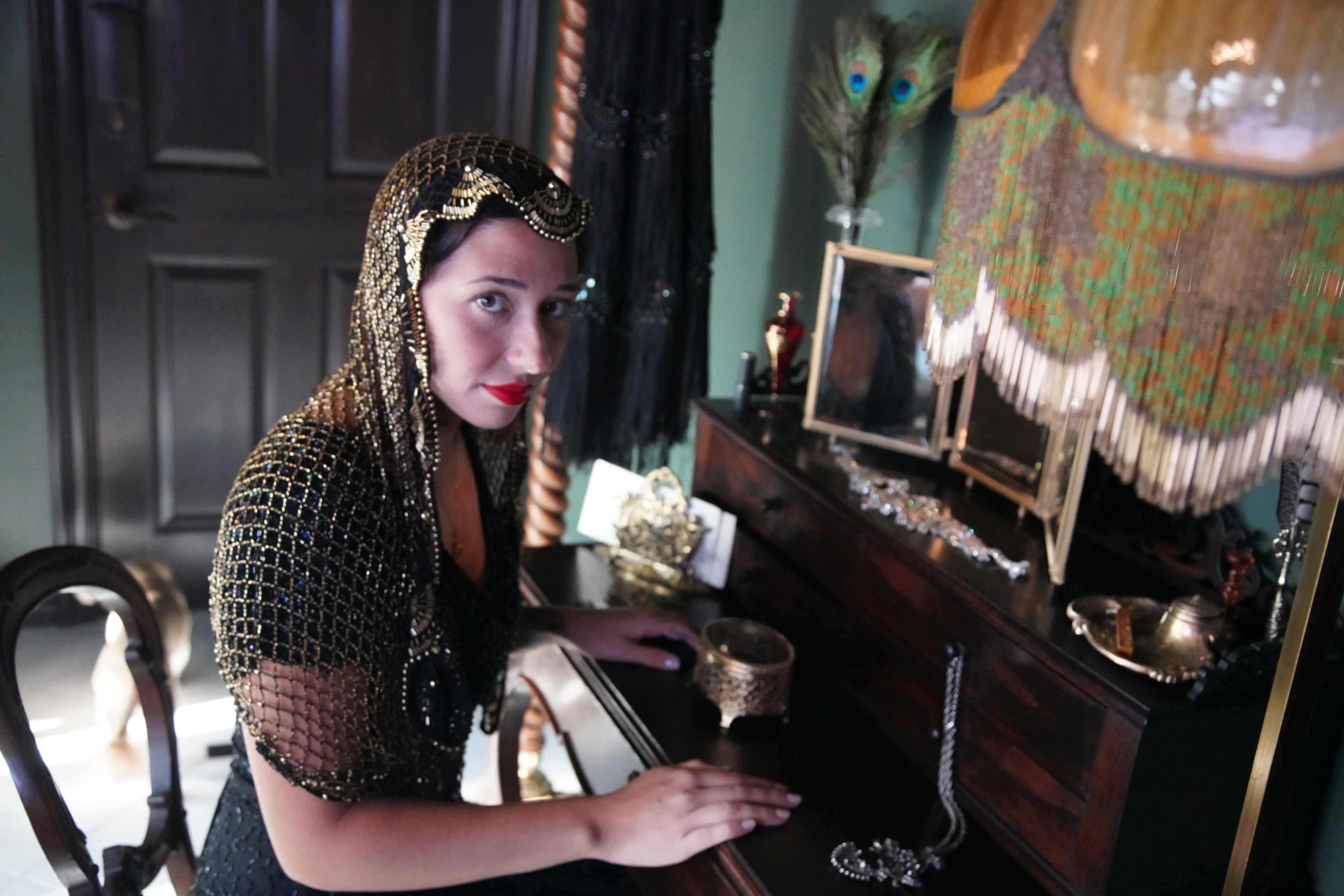 A woman in a decorative headdress sits at a vintage dressing table surrounded by various accessories and decor.