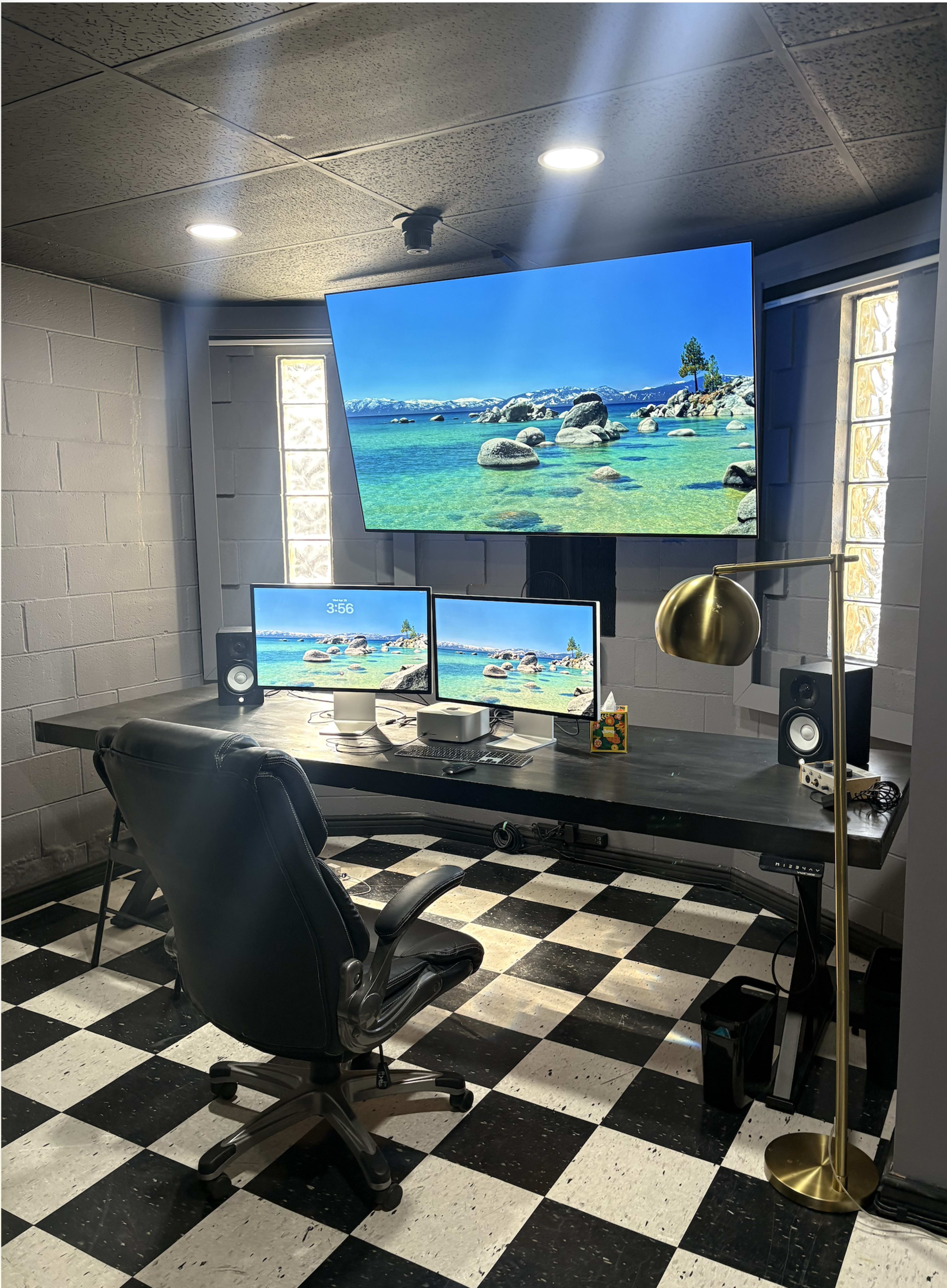 The image shows a modern home office setup featuring a large desk with dual monitors, a black swivel chair, speakers, and a decorative lamp, all positioned against a checkerboard floor and a wall with windows.