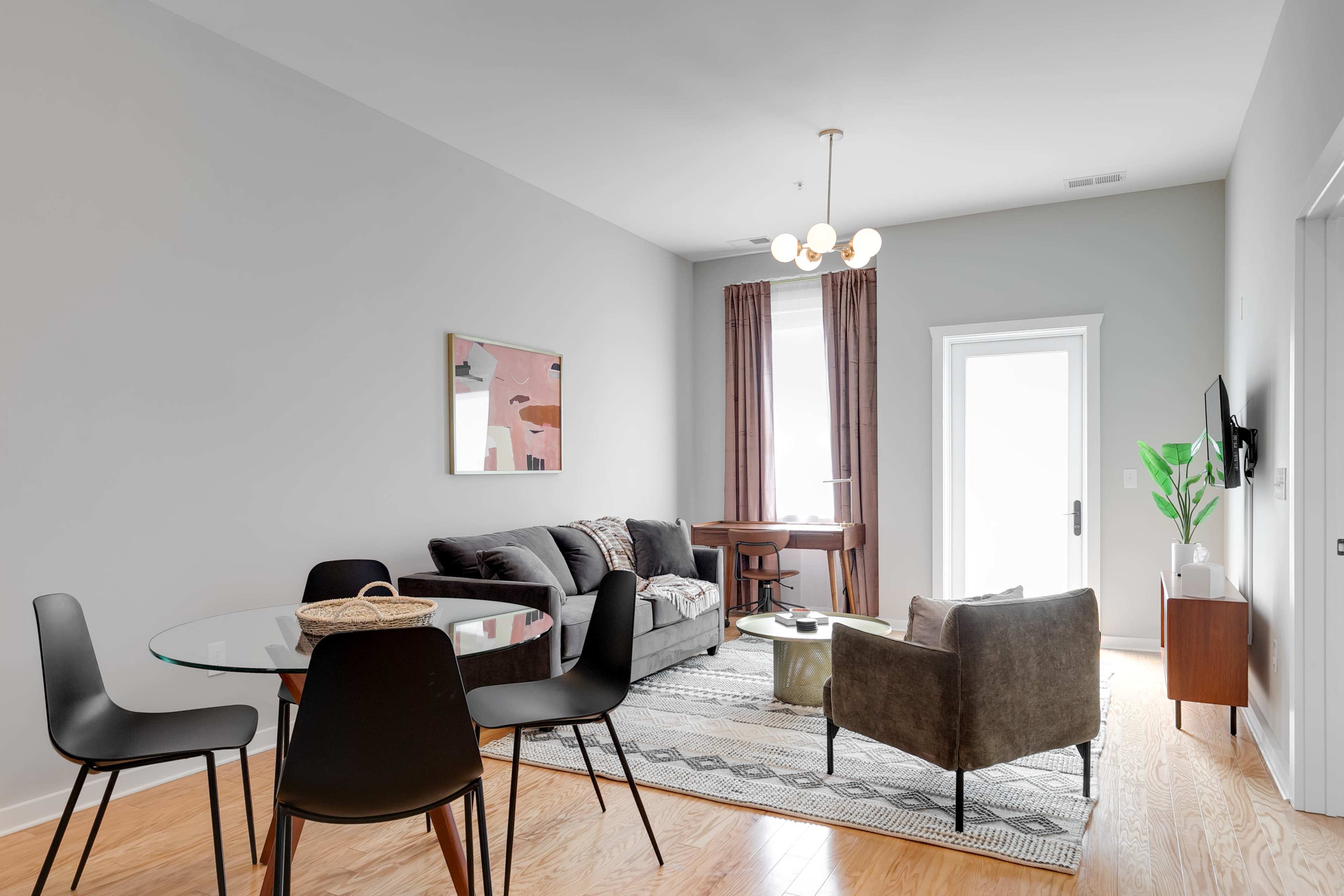 A modern living space featuring a gray sofa, a glass dining table with four black chairs, and a wooden desk near a door leading outside.