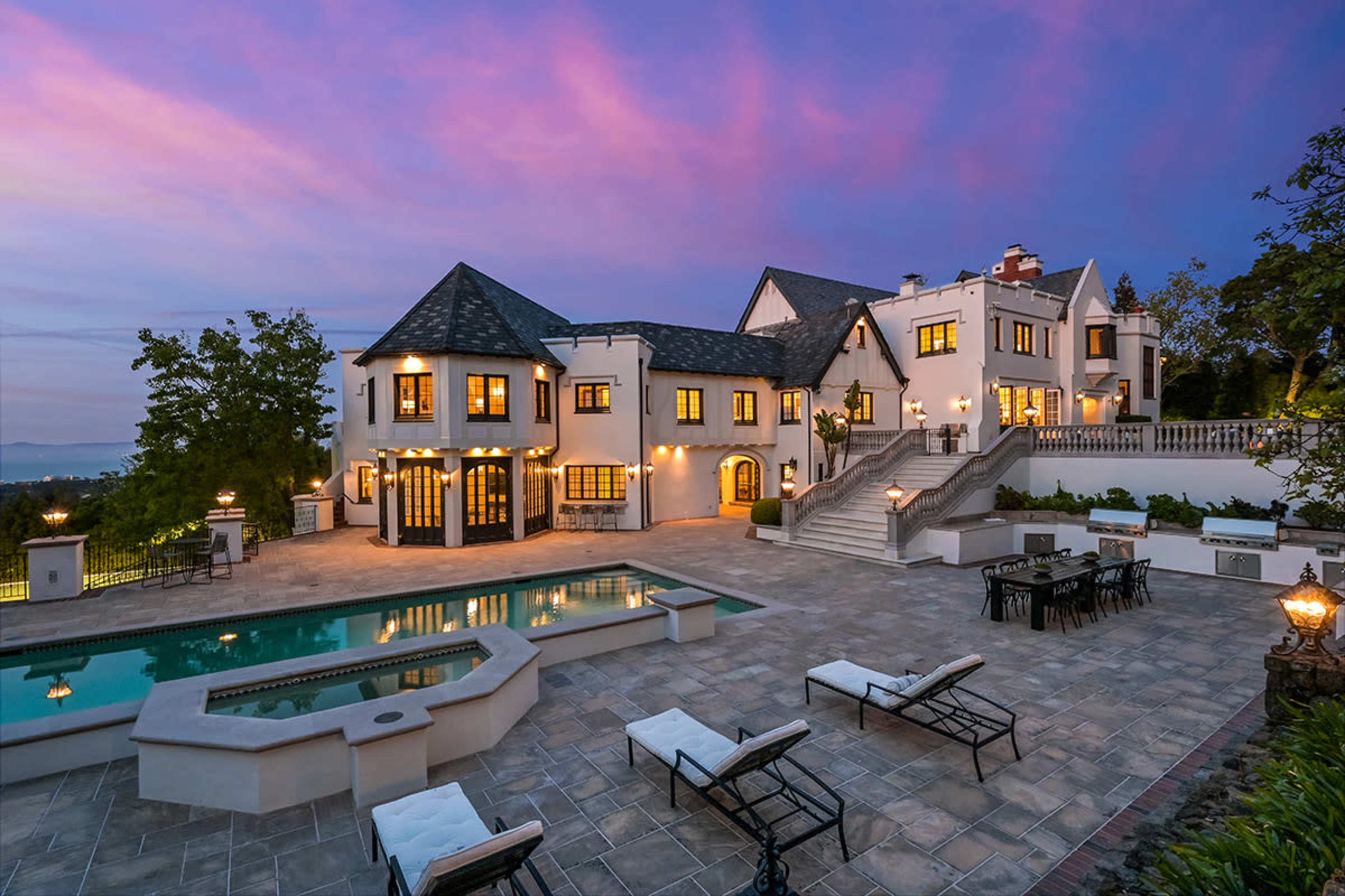 A large white mansion with multiple gabled roofs is illuminated at dusk, featuring a spacious patio with lounge chairs and a pool surrounded by landscaped gardens.