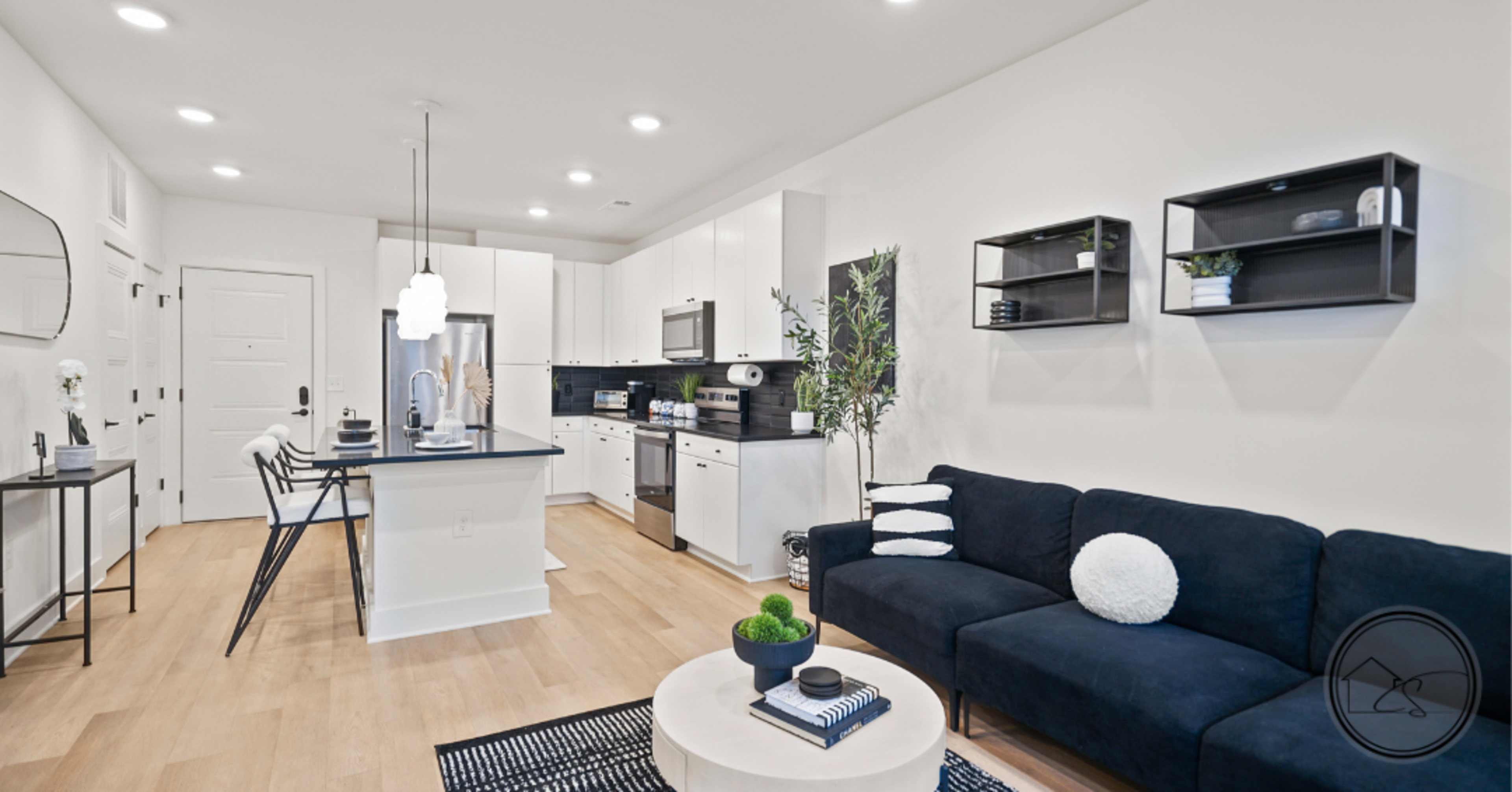 The image shows a modern, open-concept living space featuring a kitchen with white cabinetry, a black island, and a living area with a dark sofa and round coffee table.