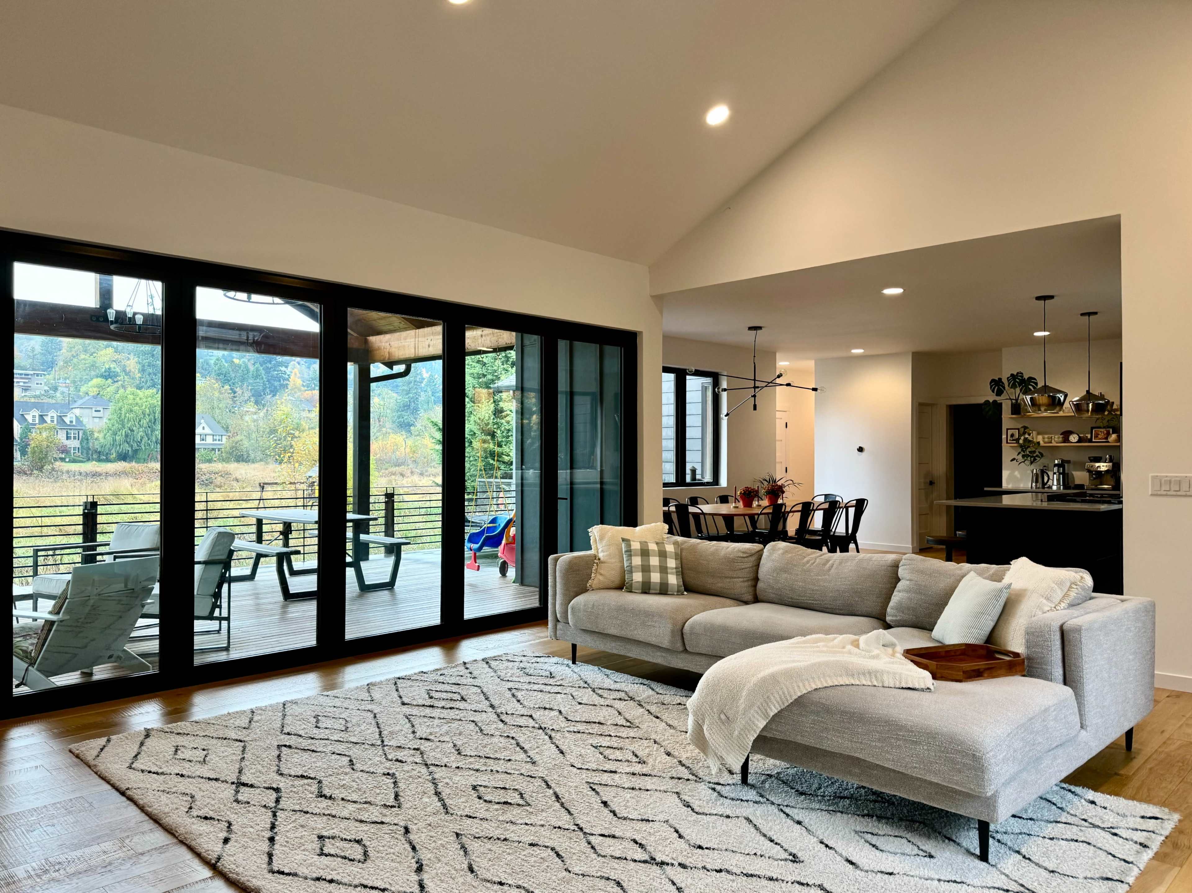 A bright and modern living room with a light-colored sofa, a patterned rug, large sliding glass doors opening to a deck, and an adjacent dining area and kitchen.