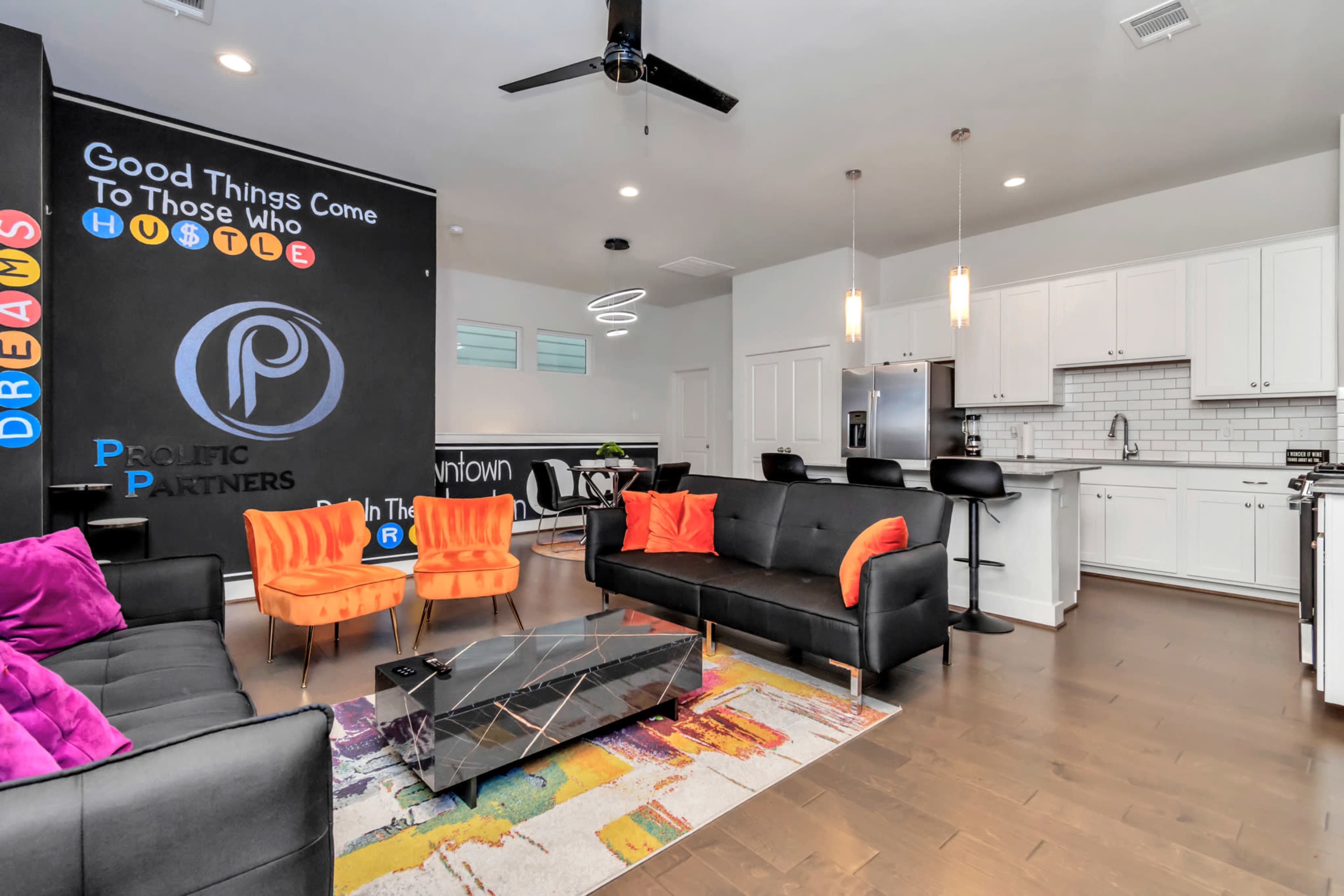 A modern living space featuring a black sofa, orange chairs, a colorful rug, and a kitchen area with white cabinetry and stainless steel appliances.