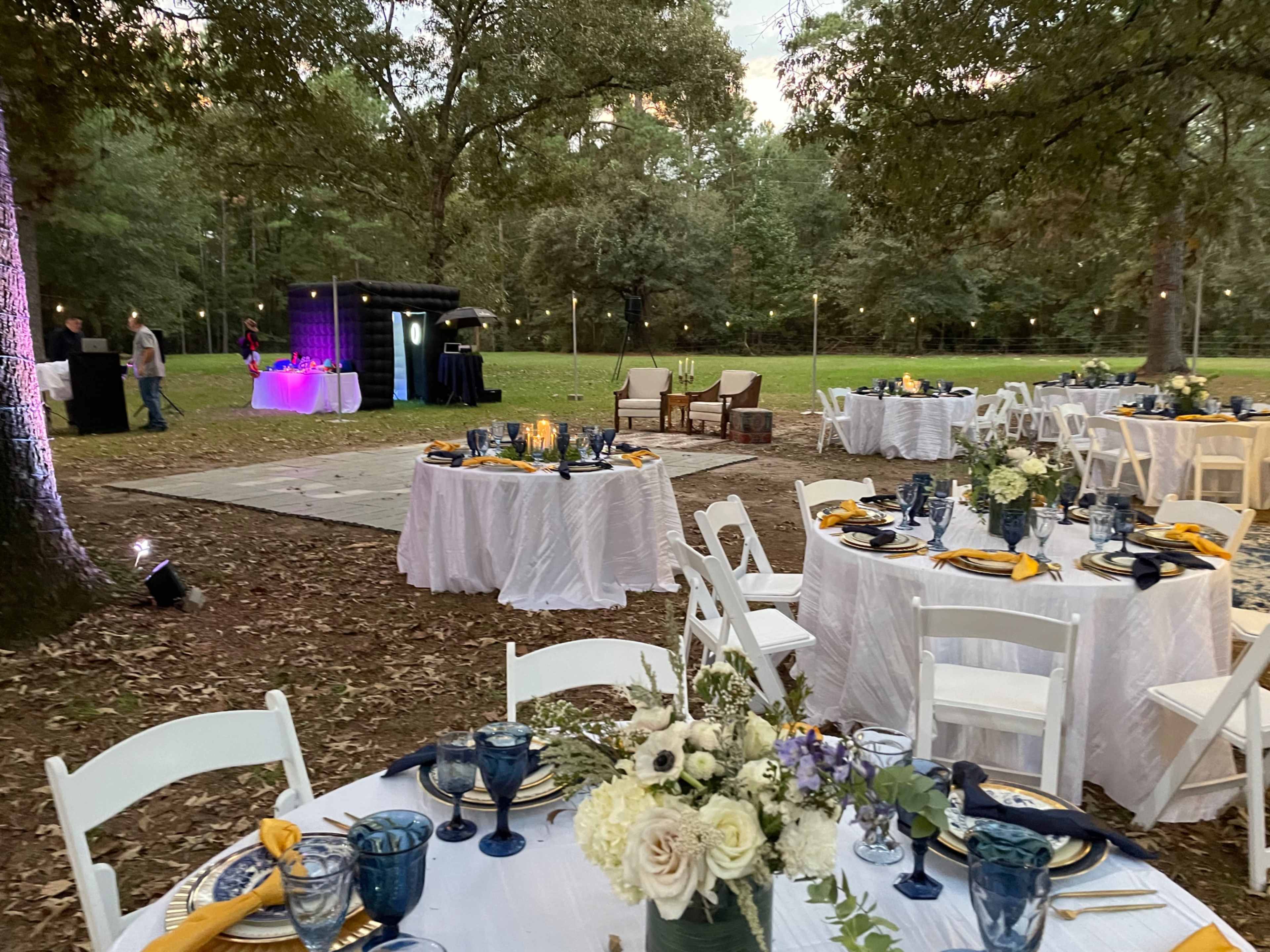 An outdoor event setup with round tables dressed in white linens, adorned with blue glassware and yellow napkins, surrounded by trees and a small stage area with lighting.