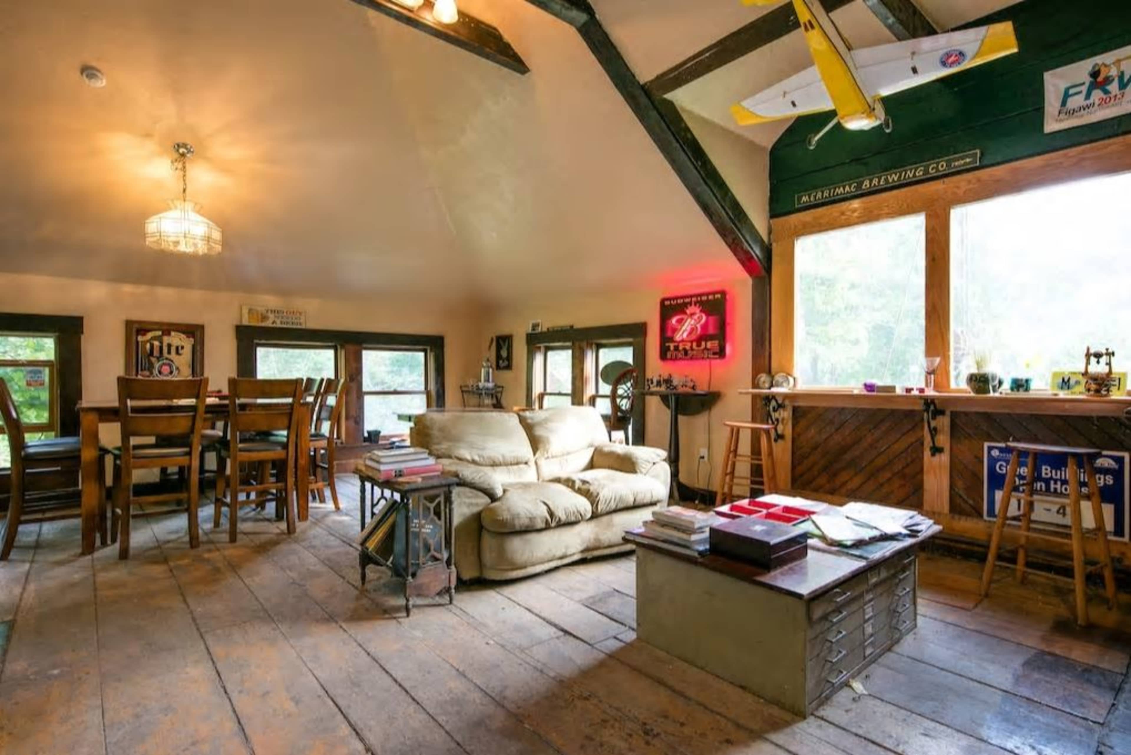 A cozy, rustic living space featuring a large couch, wooden dining table, and a bar area with windows overlooking a wooded view.