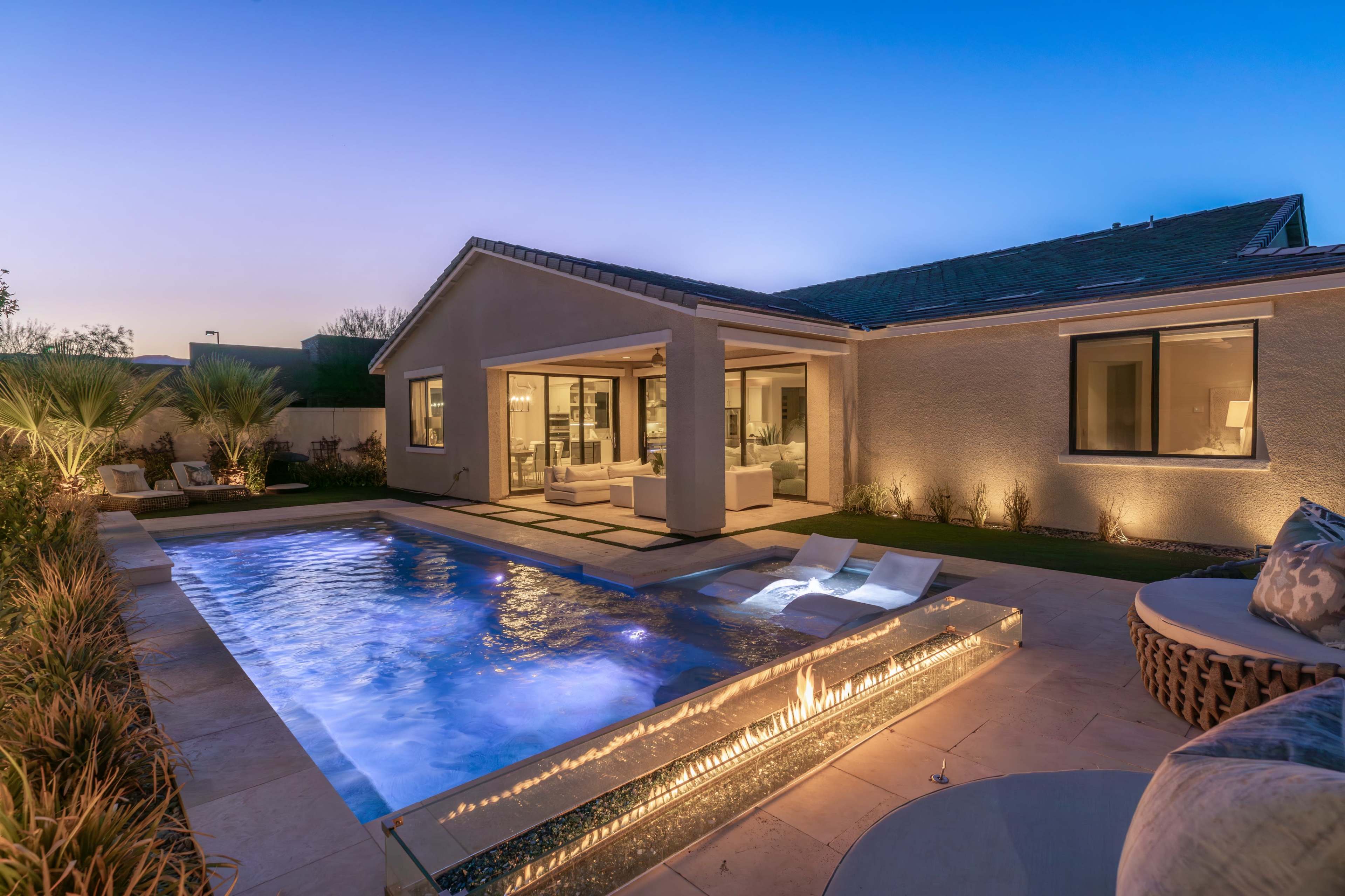 A modern backyard features a swimming pool with a built-in fire feature adjacent to a patio area in twilight.