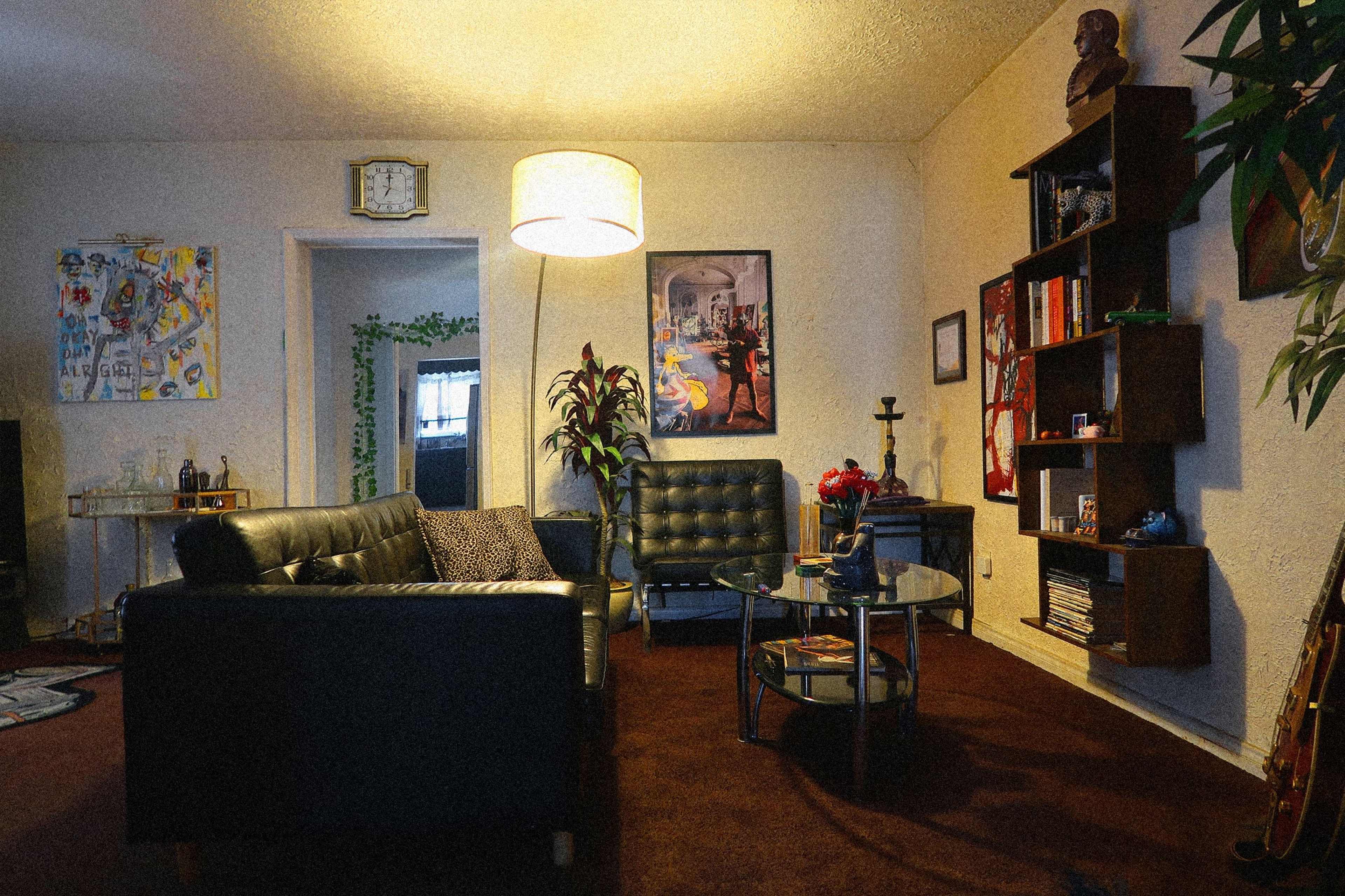 The image shows a cozy living room featuring a black leather sofa, a round glass coffee table, and various artworks on the walls, along with a lamp and a bookshelf.
