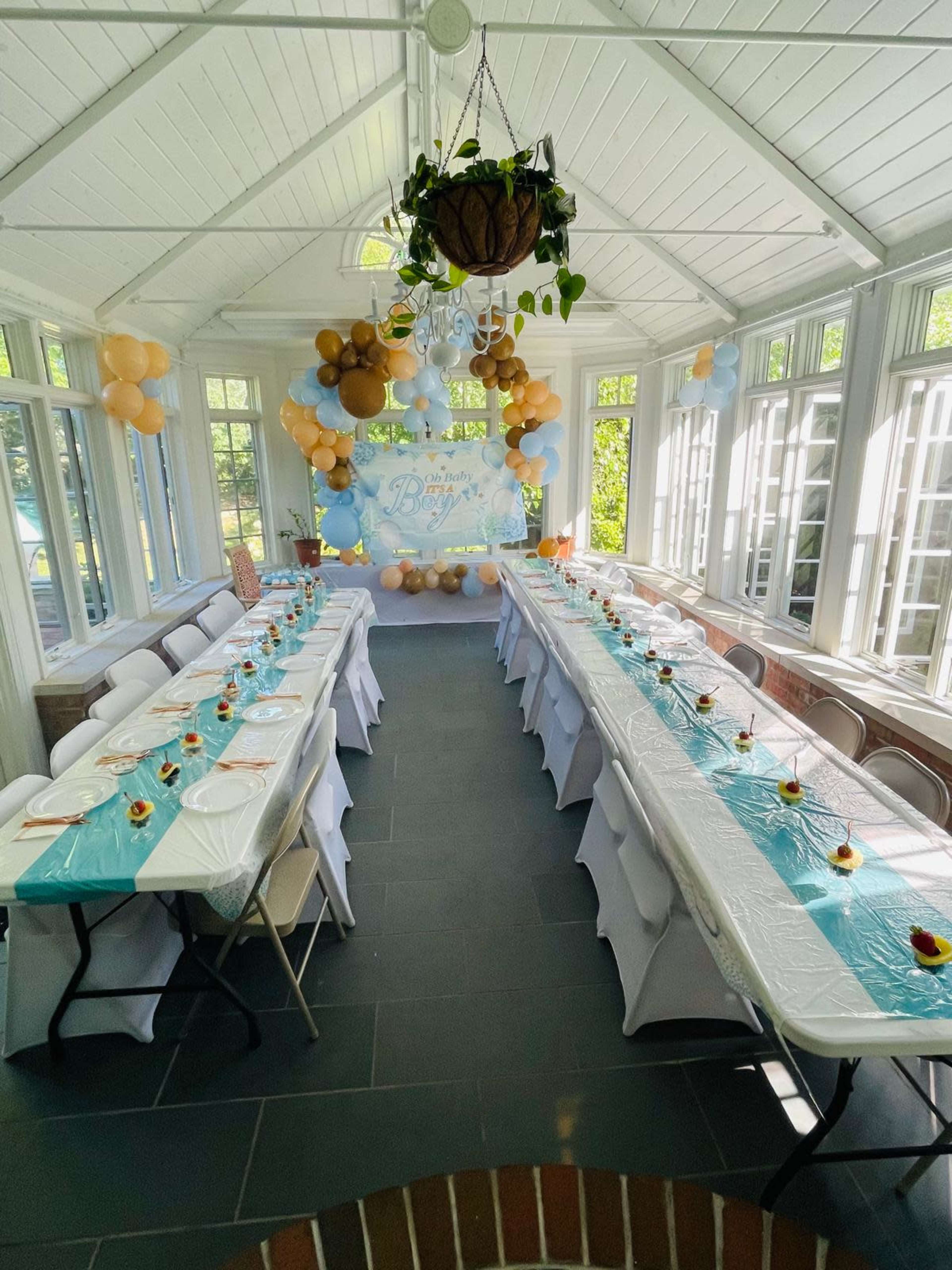 The image shows a brightly lit indoor space set up for a celebration, featuring two long tables covered in white tablecloths with blue accents and decorated with small desserts at each place setting, alongside balloons in gold and blue.