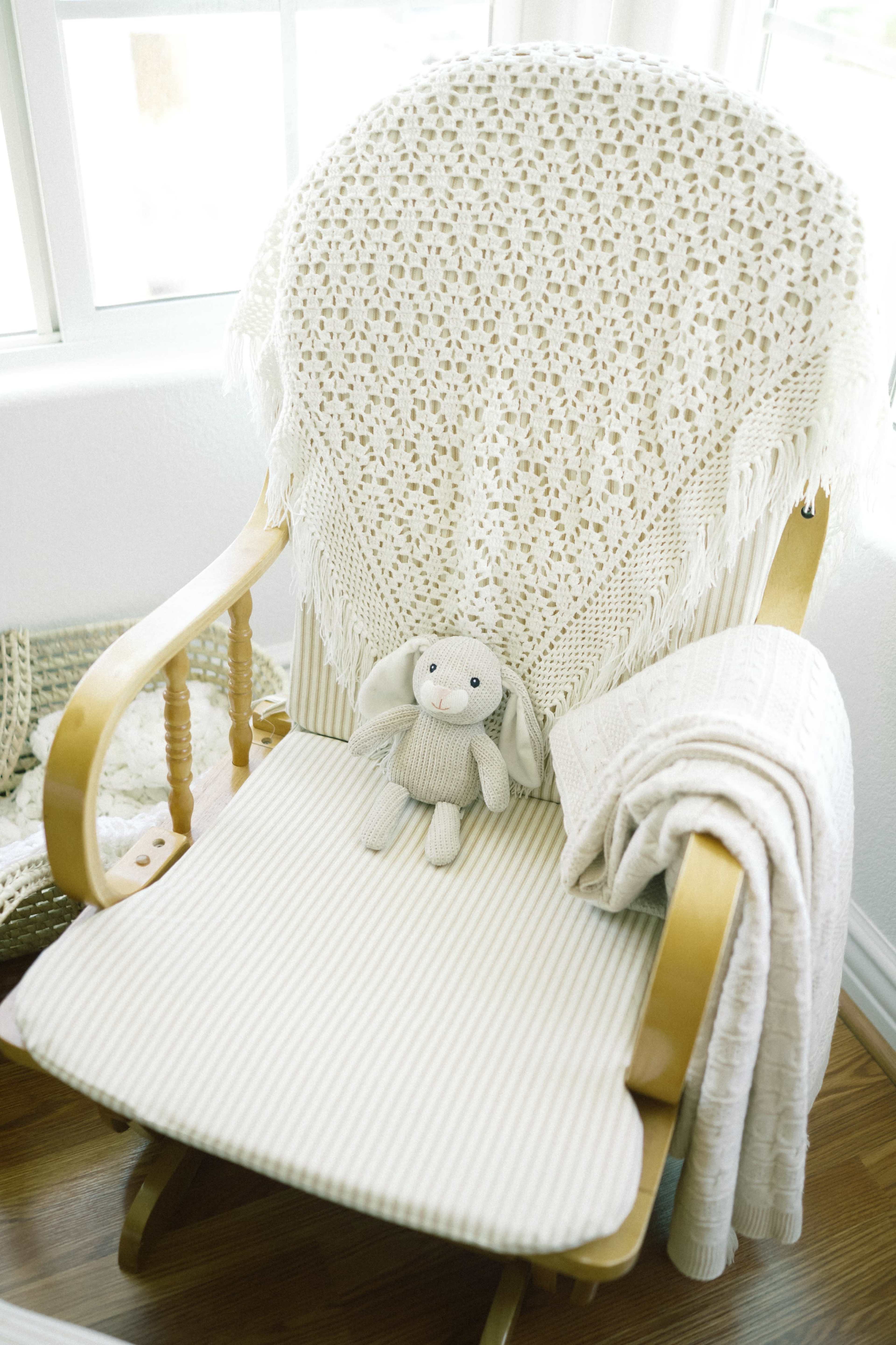 A rocking chair with a beige knitted blanket and a soft toy rabbit is positioned near a window.