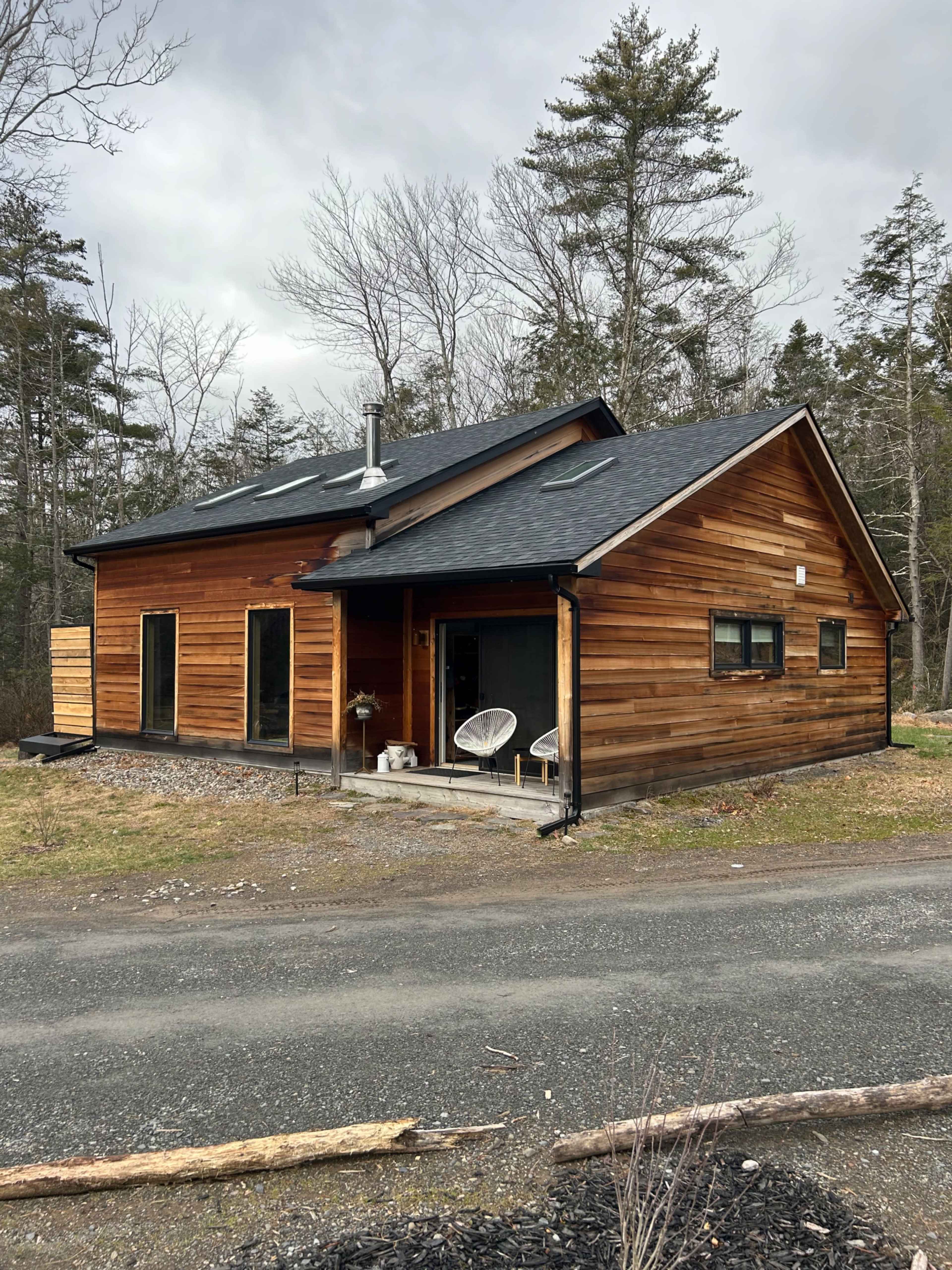 A modern wooden house with a sloped roof situated near a gravel road and surrounded by trees.