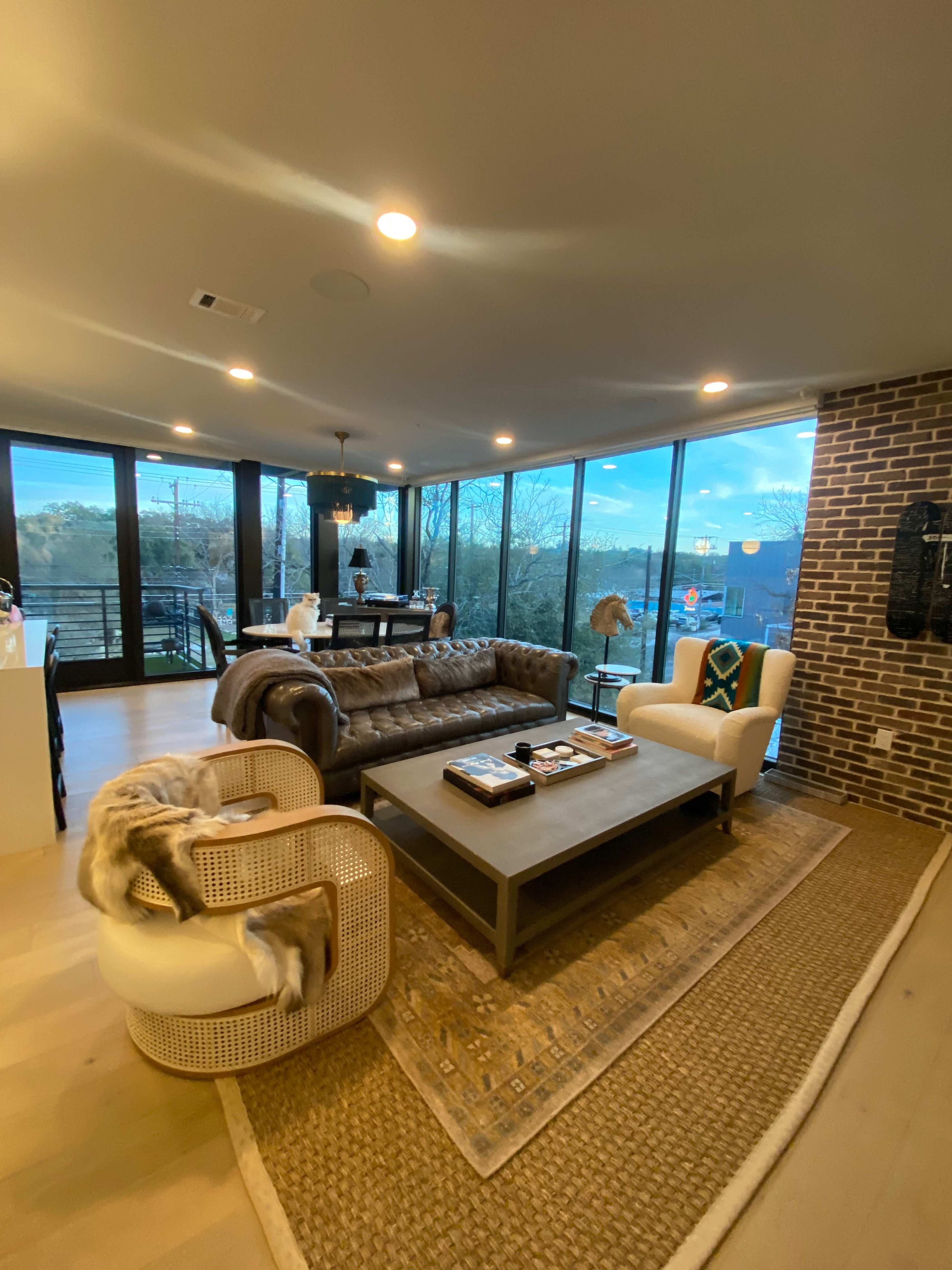The image features a modern living room with large windows, a leather sofa, two upholstered chairs, and a coffee table in front of a woven rug.