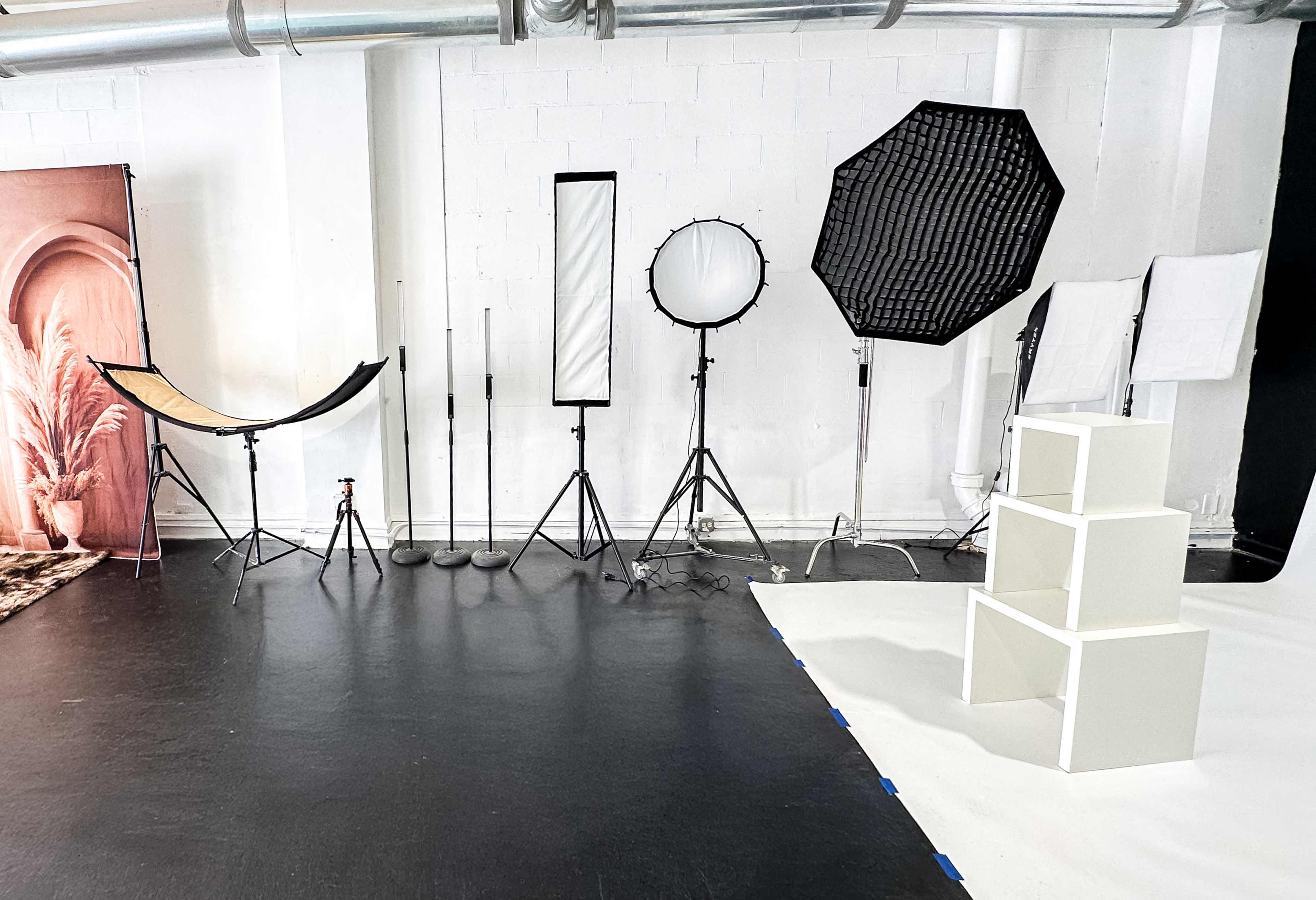 The image shows a photography studio with various lighting equipment, reflectors, and a white display stand arranged on a dark floor.