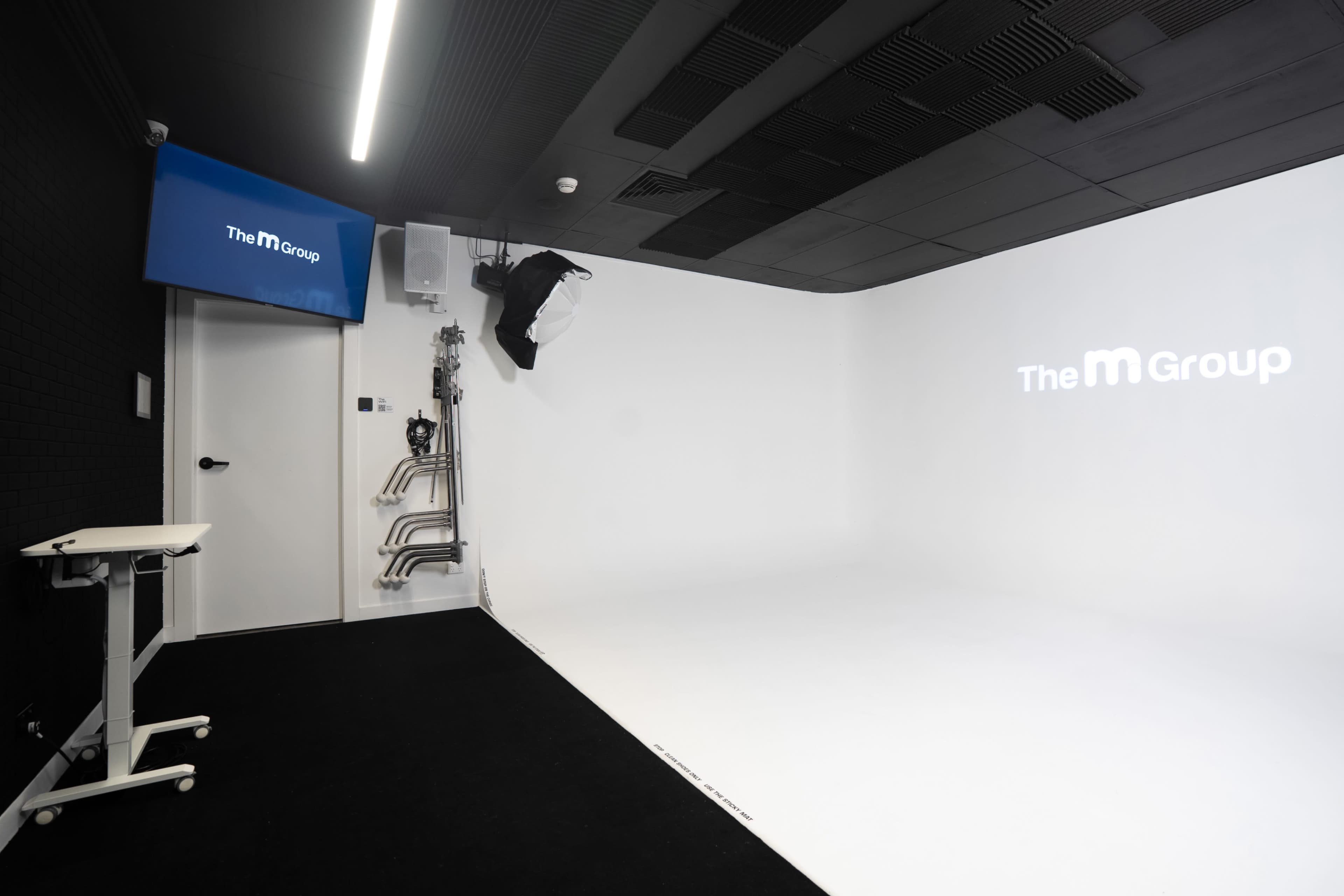 The image shows a minimalist studio space with a white backdrop, a screen displaying "The M Group," and a few studio equipment elements mounted on the wall.