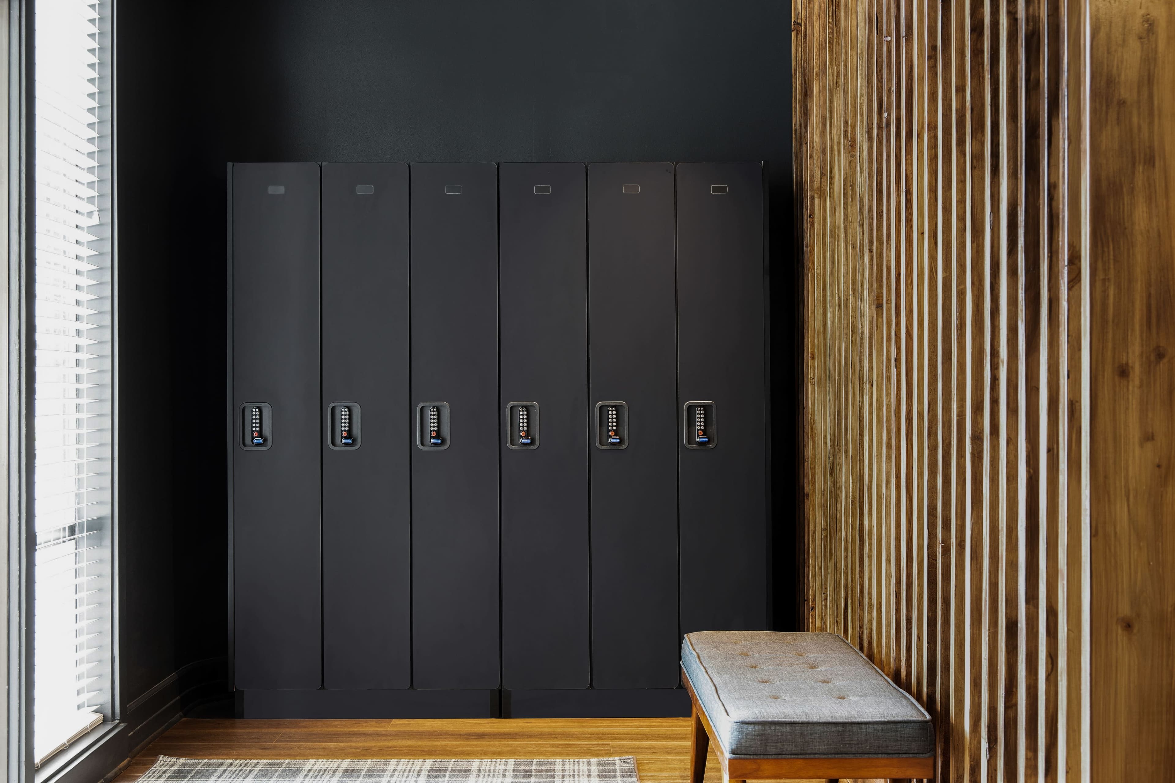 A row of six black lockers against a dark wall, with a wooden bench positioned nearby.
