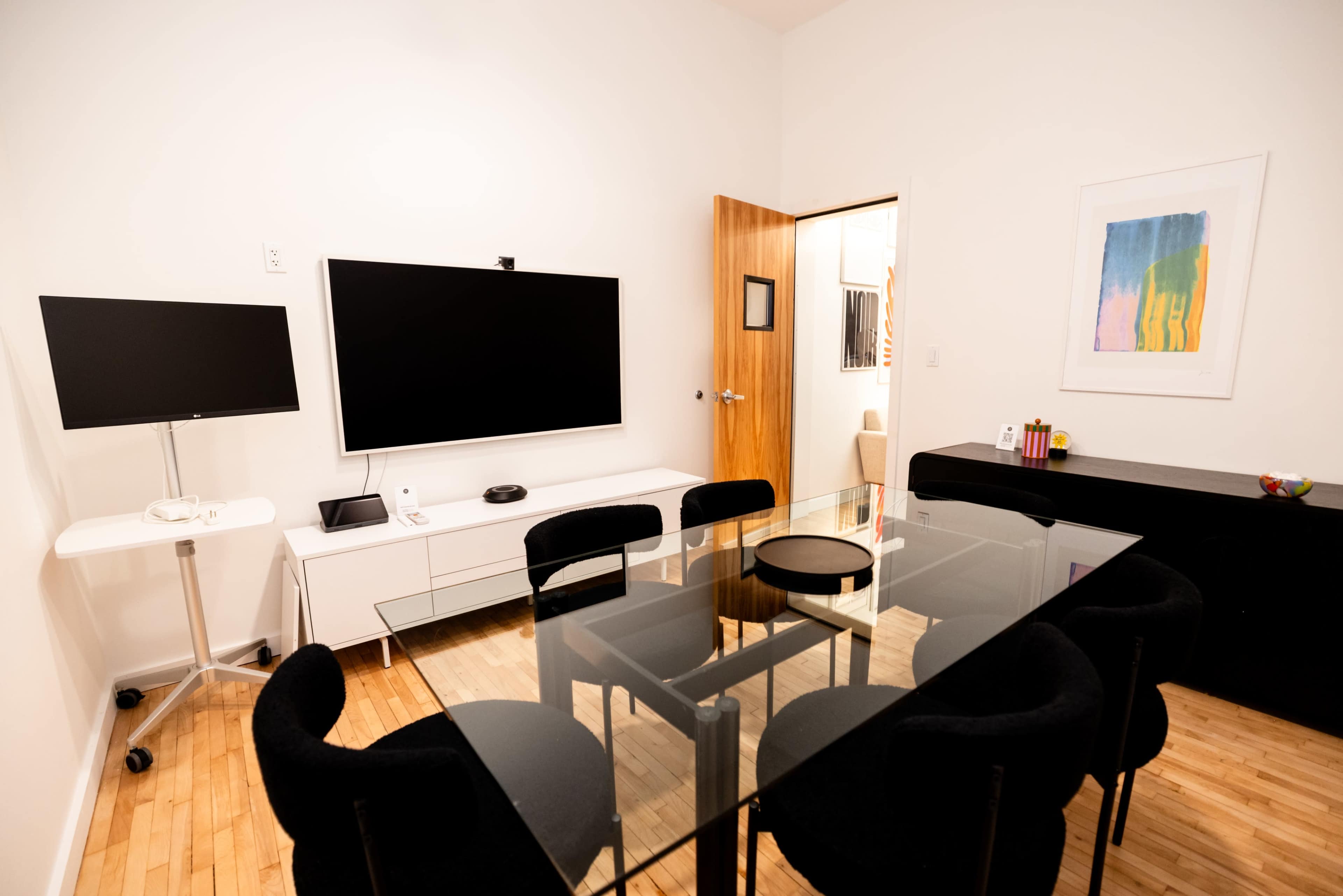 The image shows a modern office meeting room featuring a glass table, black chairs, a large television, a smaller monitor on wheels, and a wooden door leading to another space.