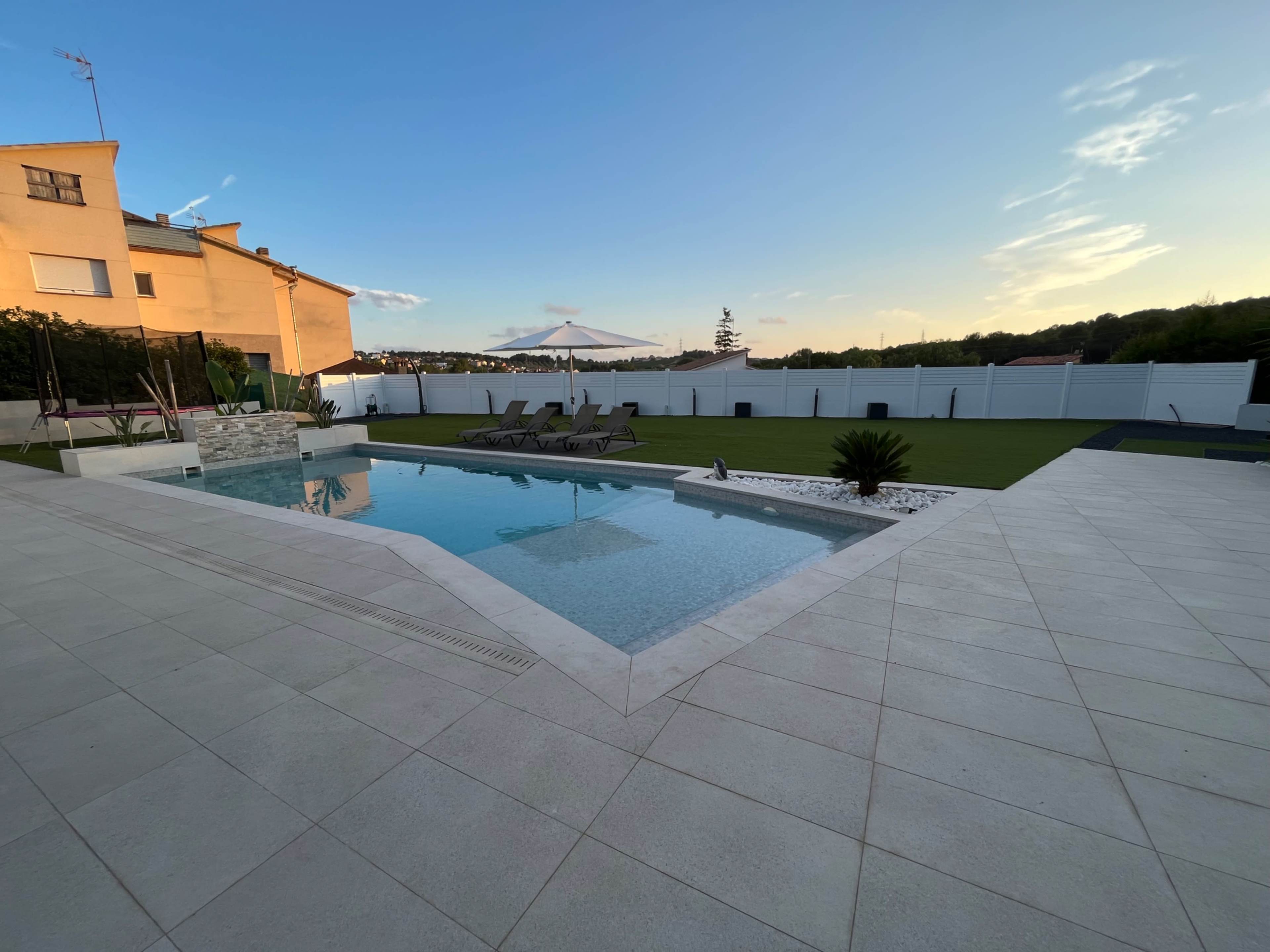 A modern backyard with a swimming pool surrounded by a paved area, loungers, and a grass lawn.